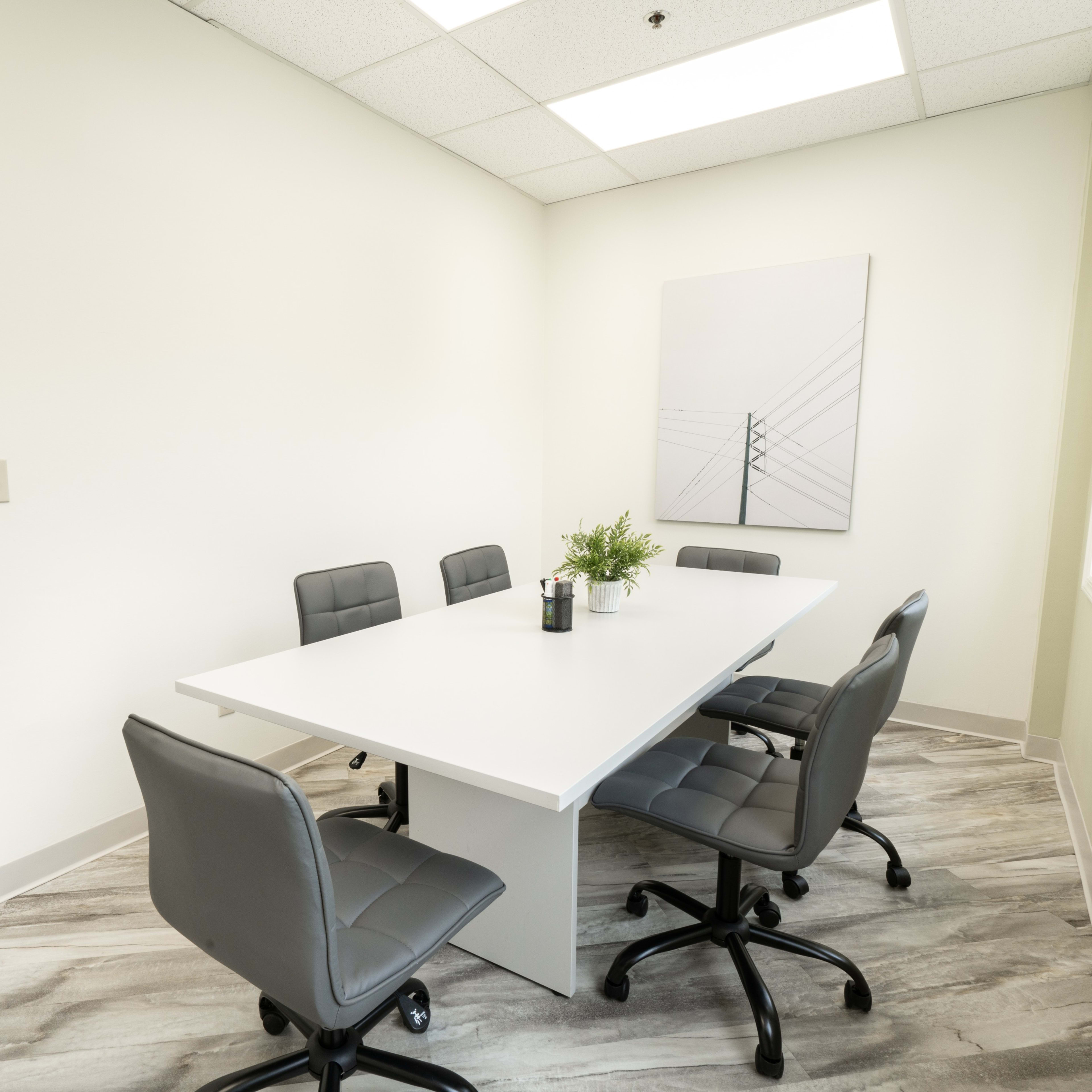 A conference room features a rectangular white table surrounded by five gray office chairs against light-colored walls.