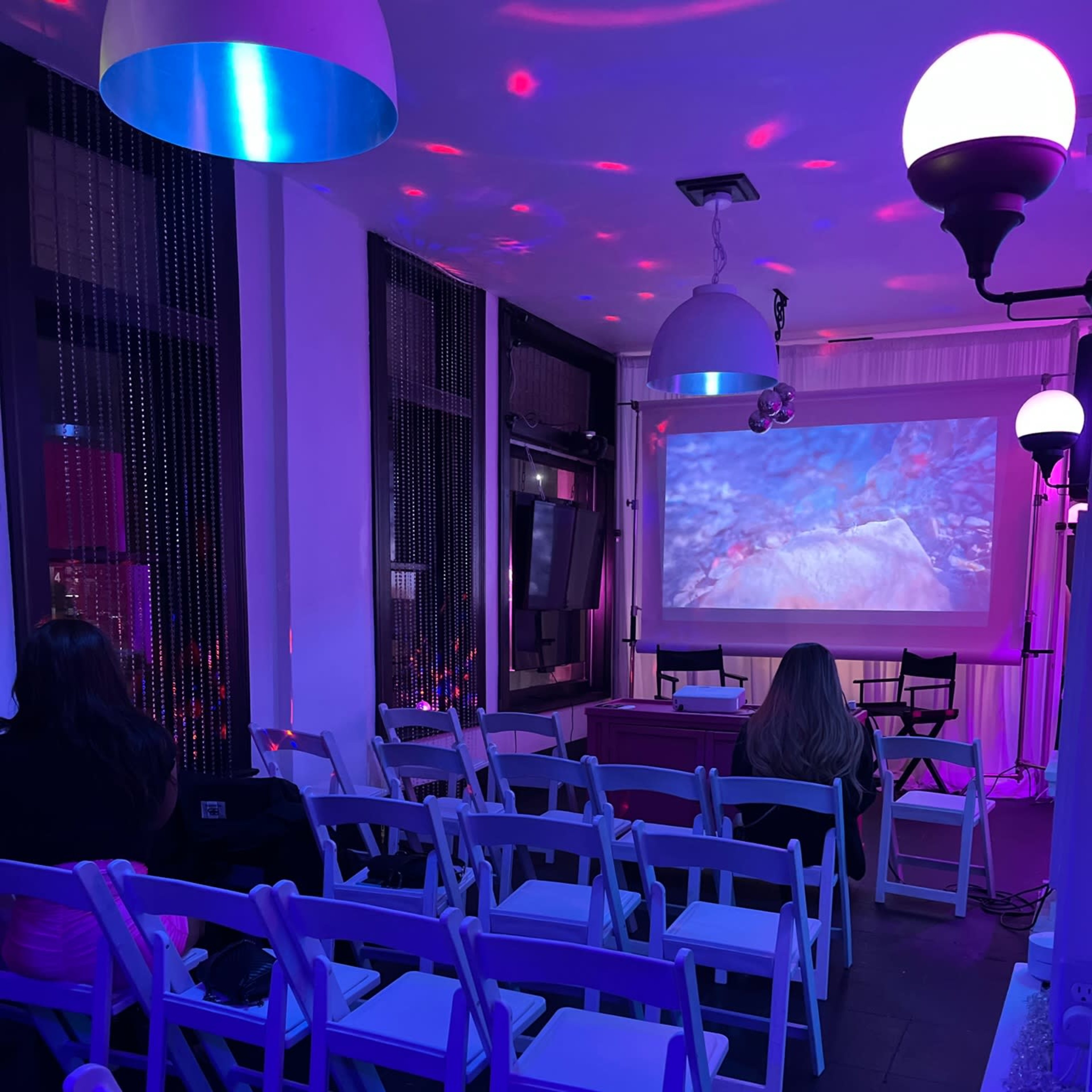 A dimly lit room set up for an event, featuring rows of white chairs facing a screen displaying a colorful projection.