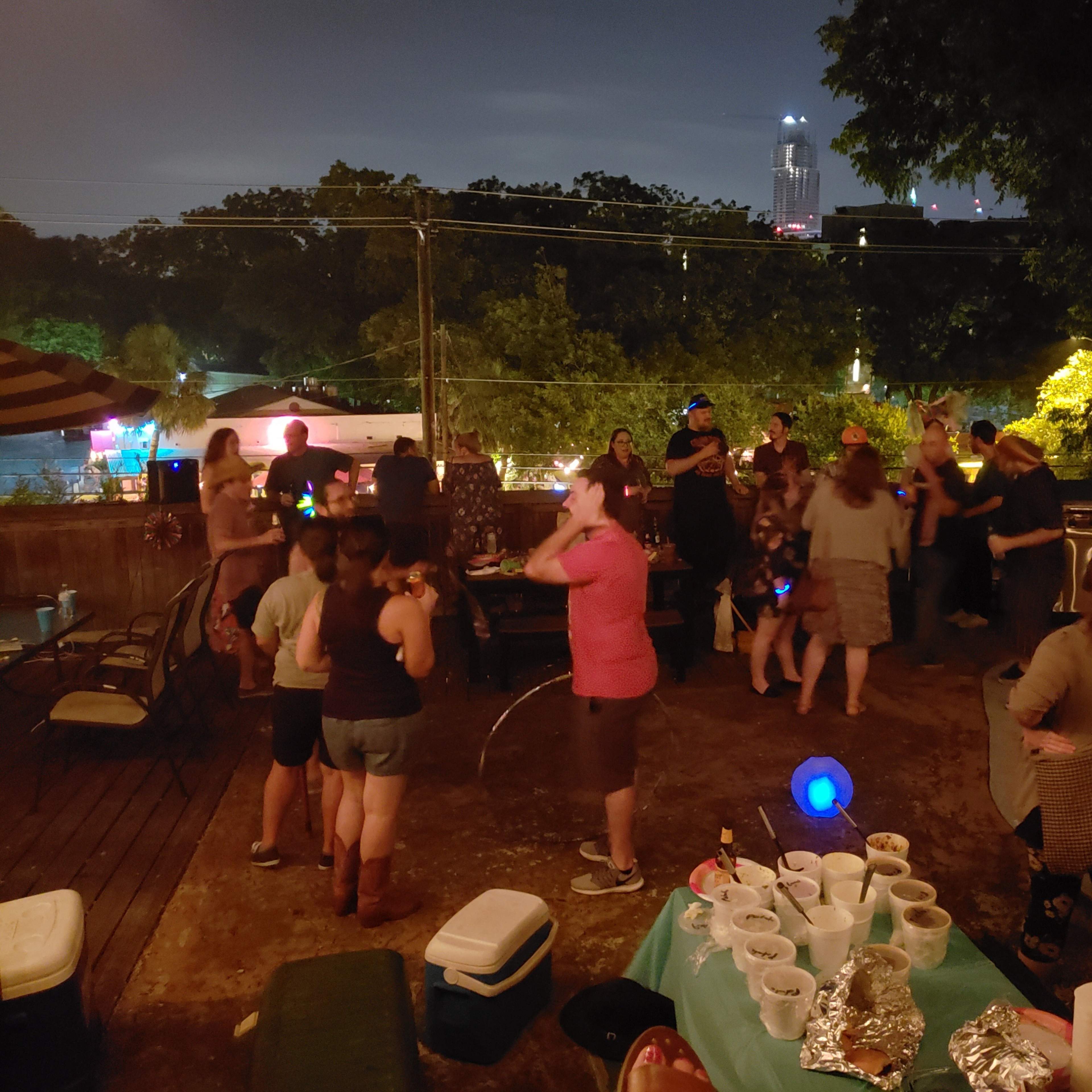 A group of people is dancing and socializing at an outdoor gathering on a deck at night, with city lights visible in the background.