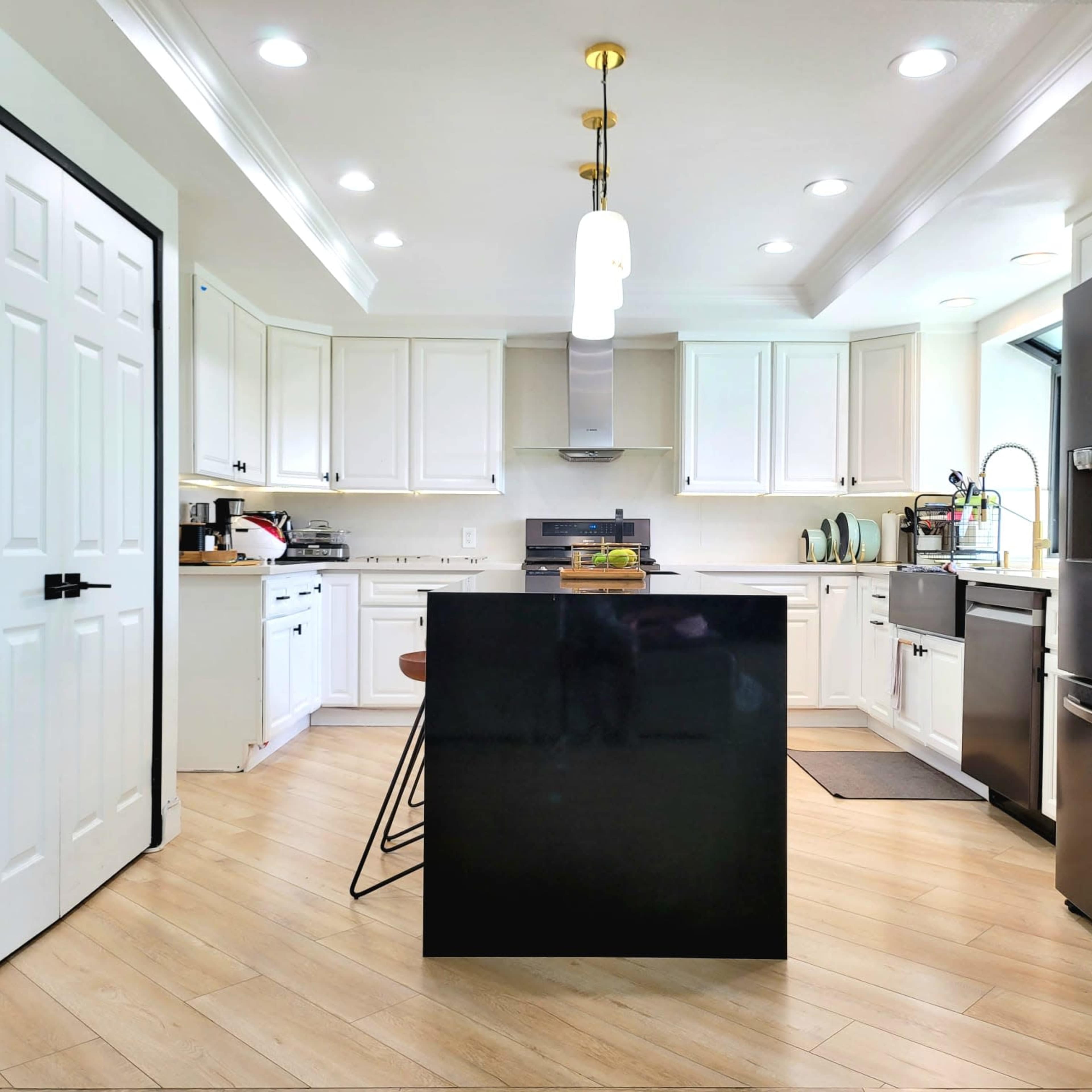 A modern kitchen with white cabinets, a black island, stainless steel appliances, and wooden flooring.