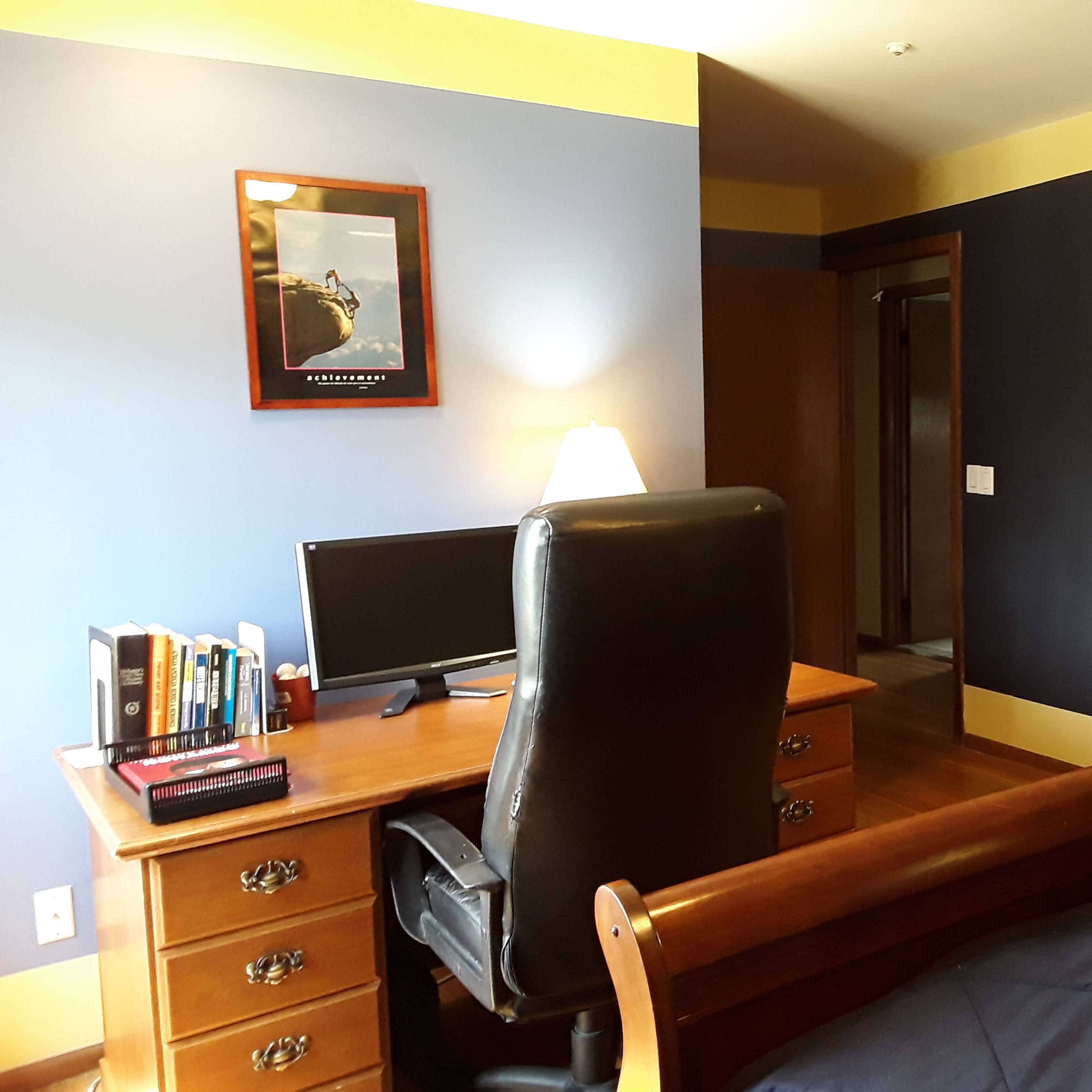 A neatly arranged bedroom featuring a wooden desk with a computer, a leather chair, and a dresser against a wall with contrasting paint colors.