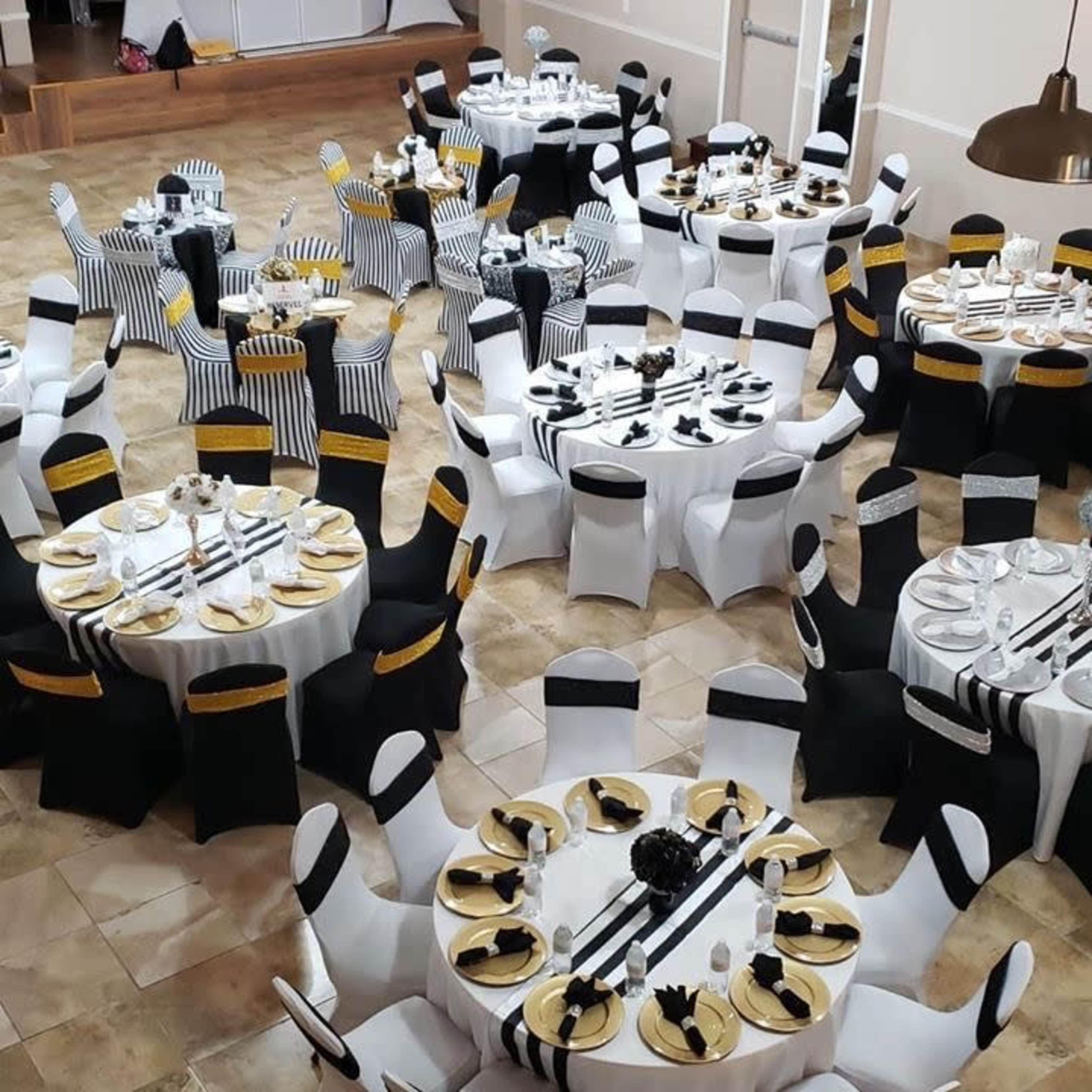 A banquet hall arranged with multiple round tables dressed in white and black tablecloths, each set with gold plates and cutlery.
