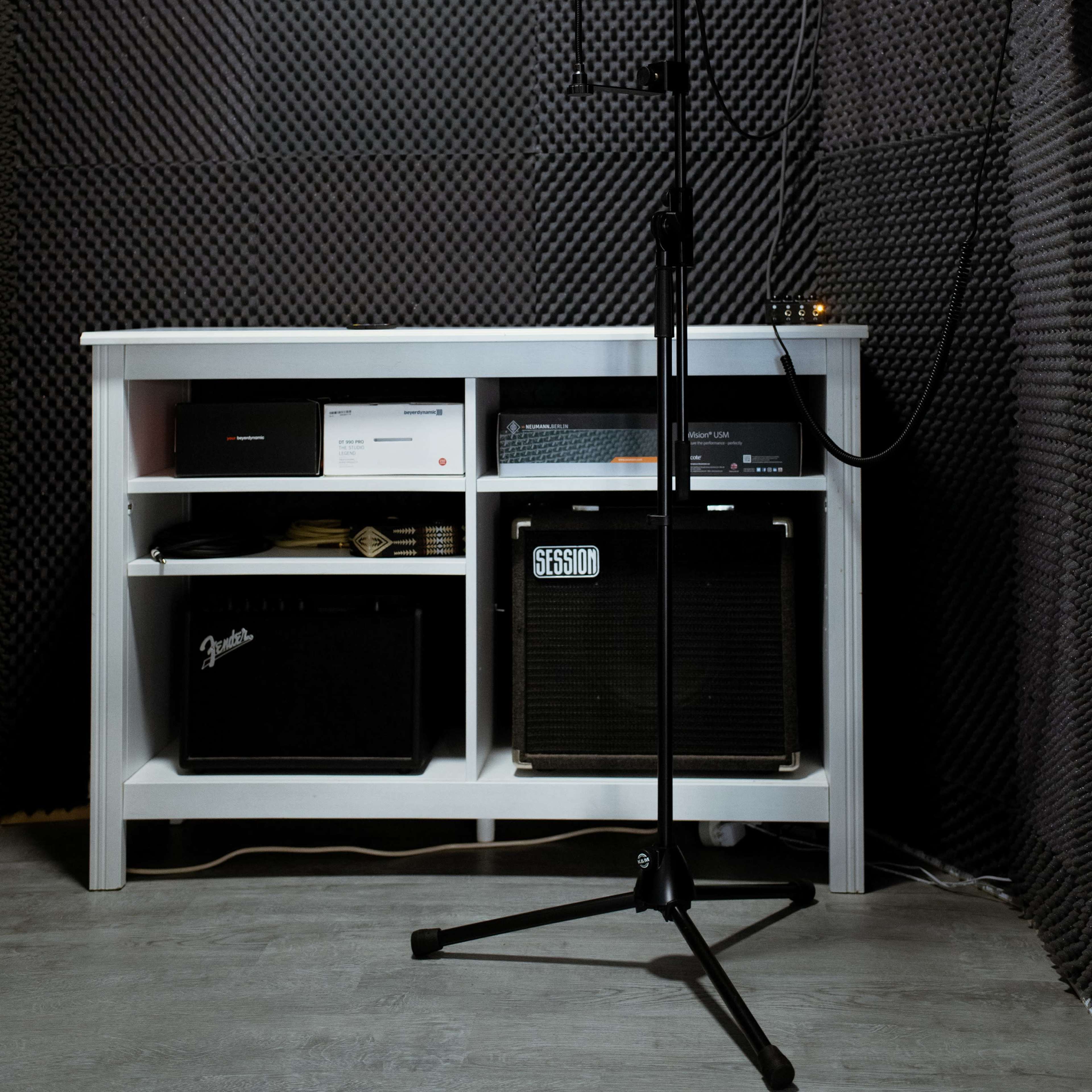 A microphone stand is positioned in front of a white shelf filled with audio equipment, all set within a soundproof room lined with black acoustic panels.