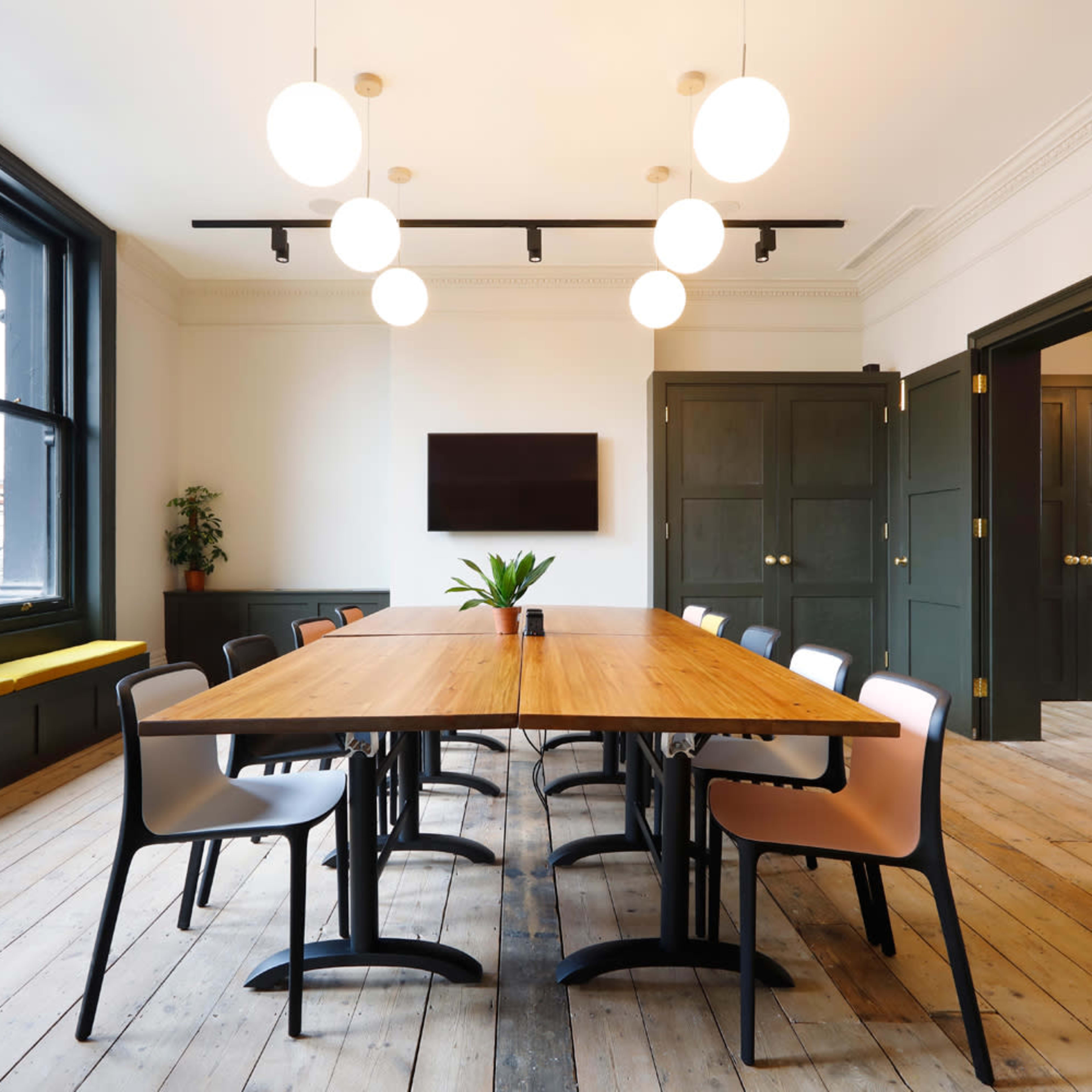 A modern conference room features a long wooden table surrounded by chairs, with large windows and minimalist lighting fixtures.