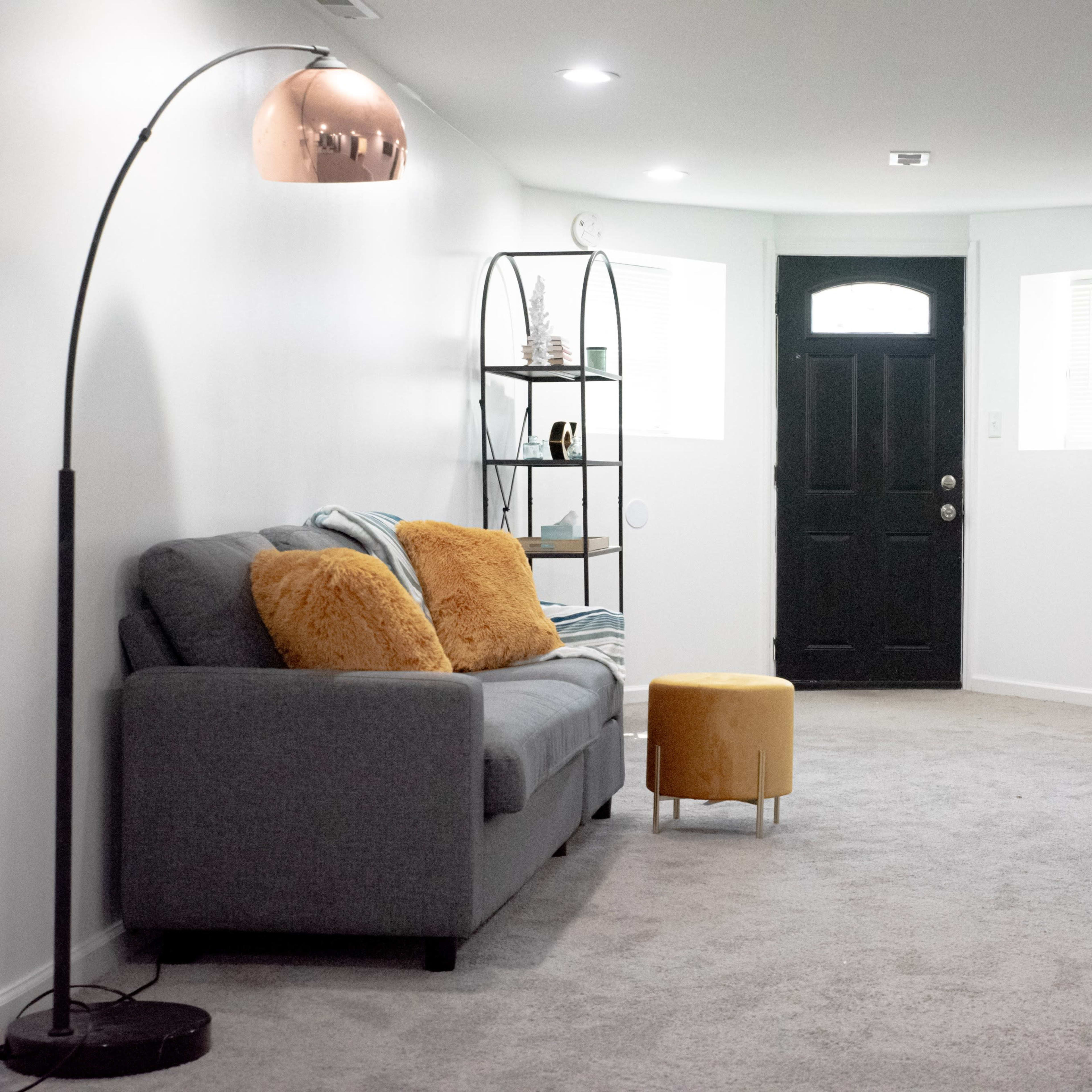A modern living space with a gray sofa, a yellow ottoman, a floor lamp, and a black door, all set against light gray walls and carpet.