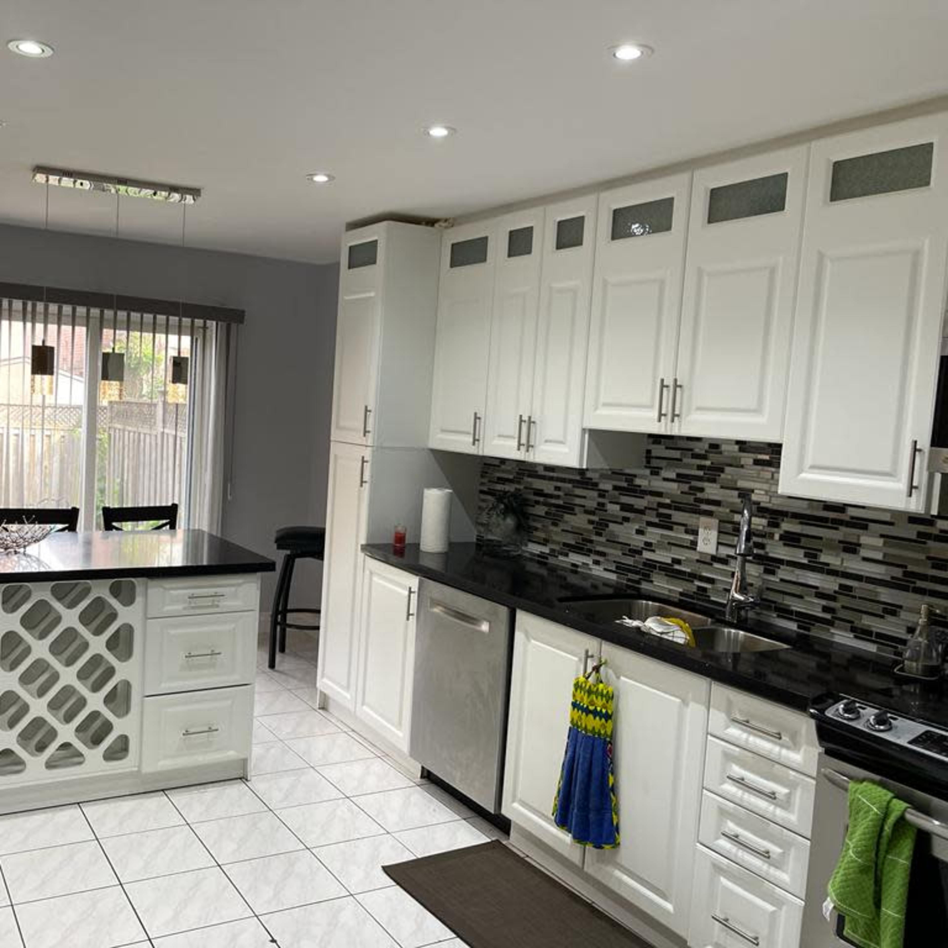 A modern kitchen with white cabinets, a black countertop, and a tiled floor, featuring a small dining area visible through an open doorway.