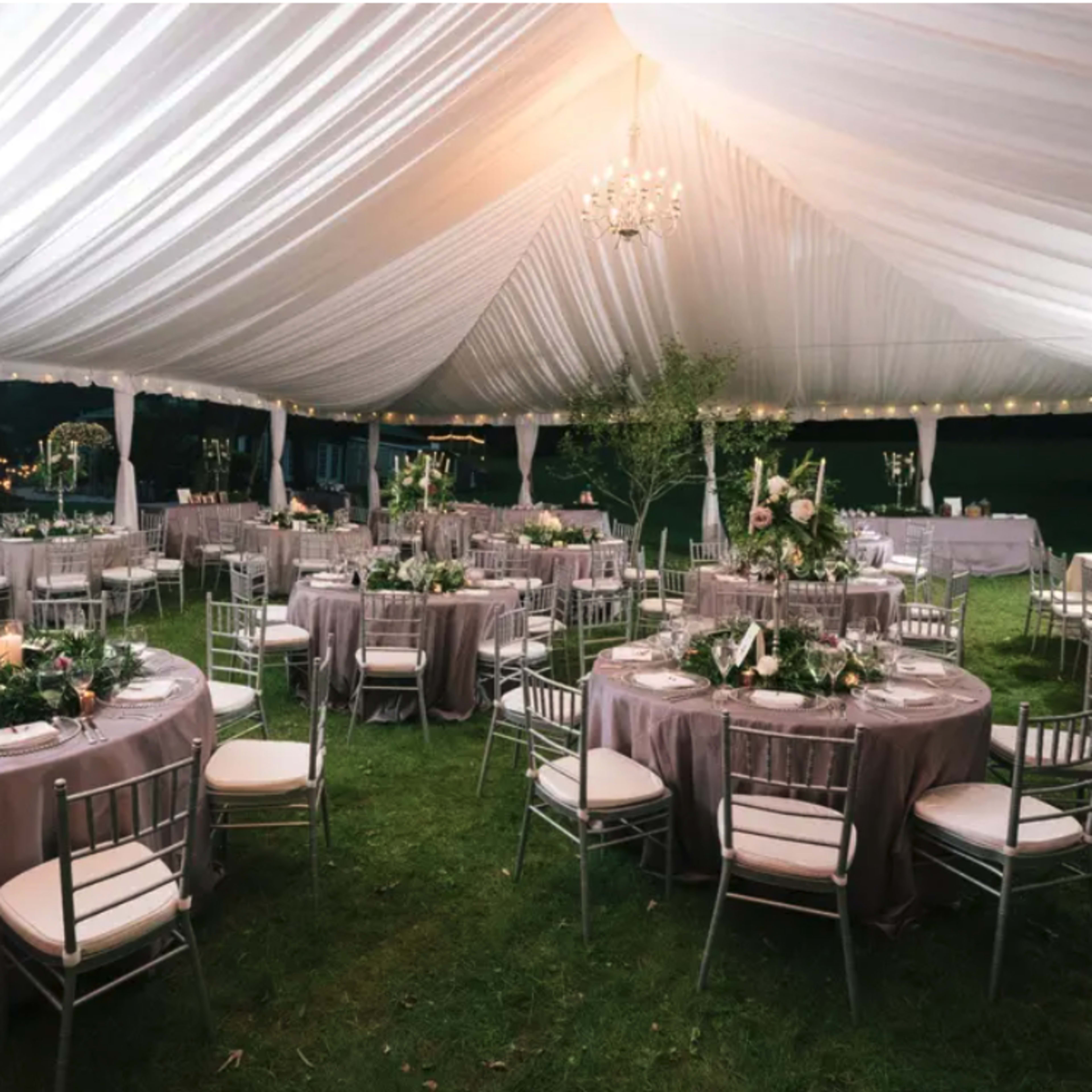 A large outdoor tent is set up for an event, featuring round tables with gray tablecloths and silver chairs, all decorated with floral centerpieces and candles.