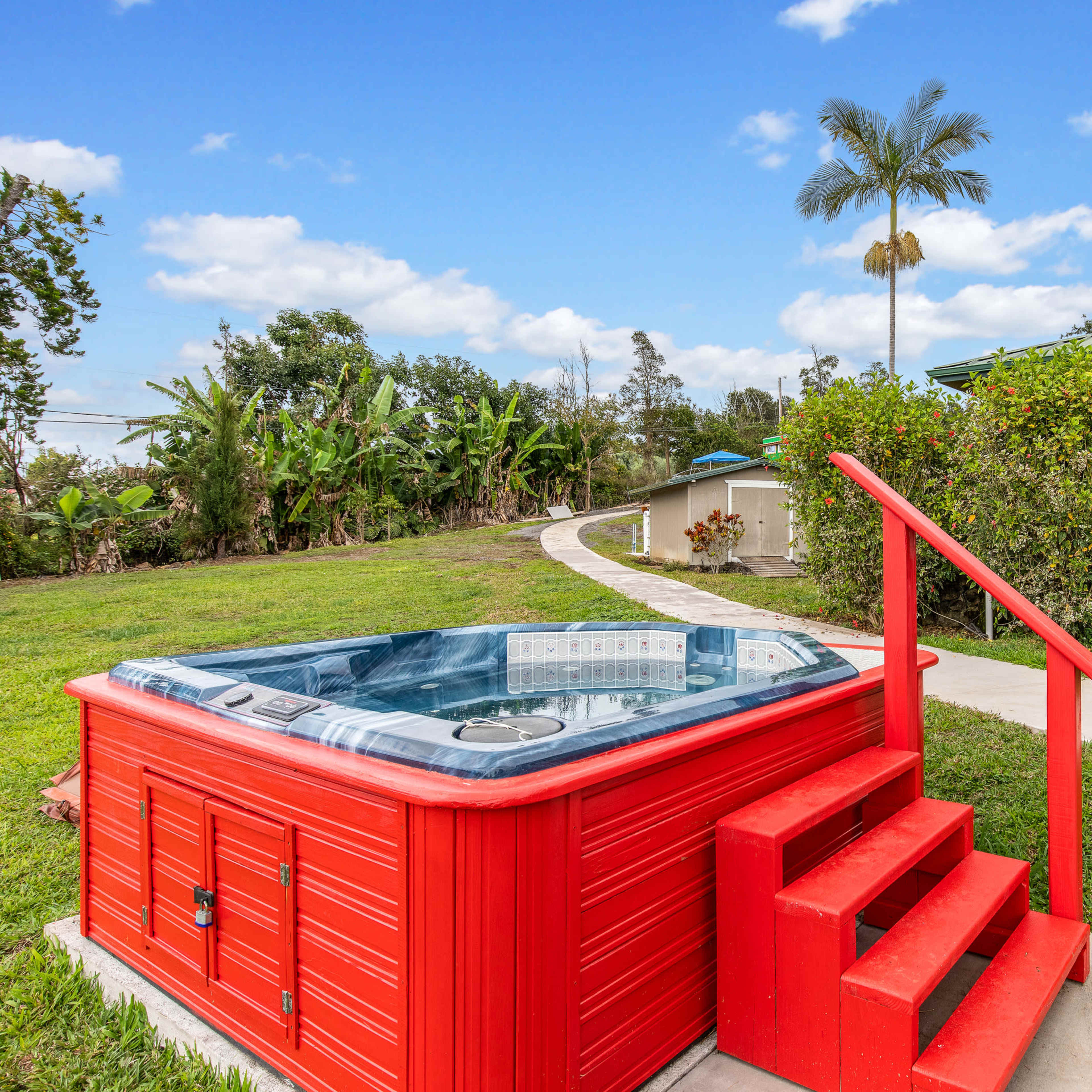 A red hot tub with steps is located beside a pathway that leads through green grass and shrubs.