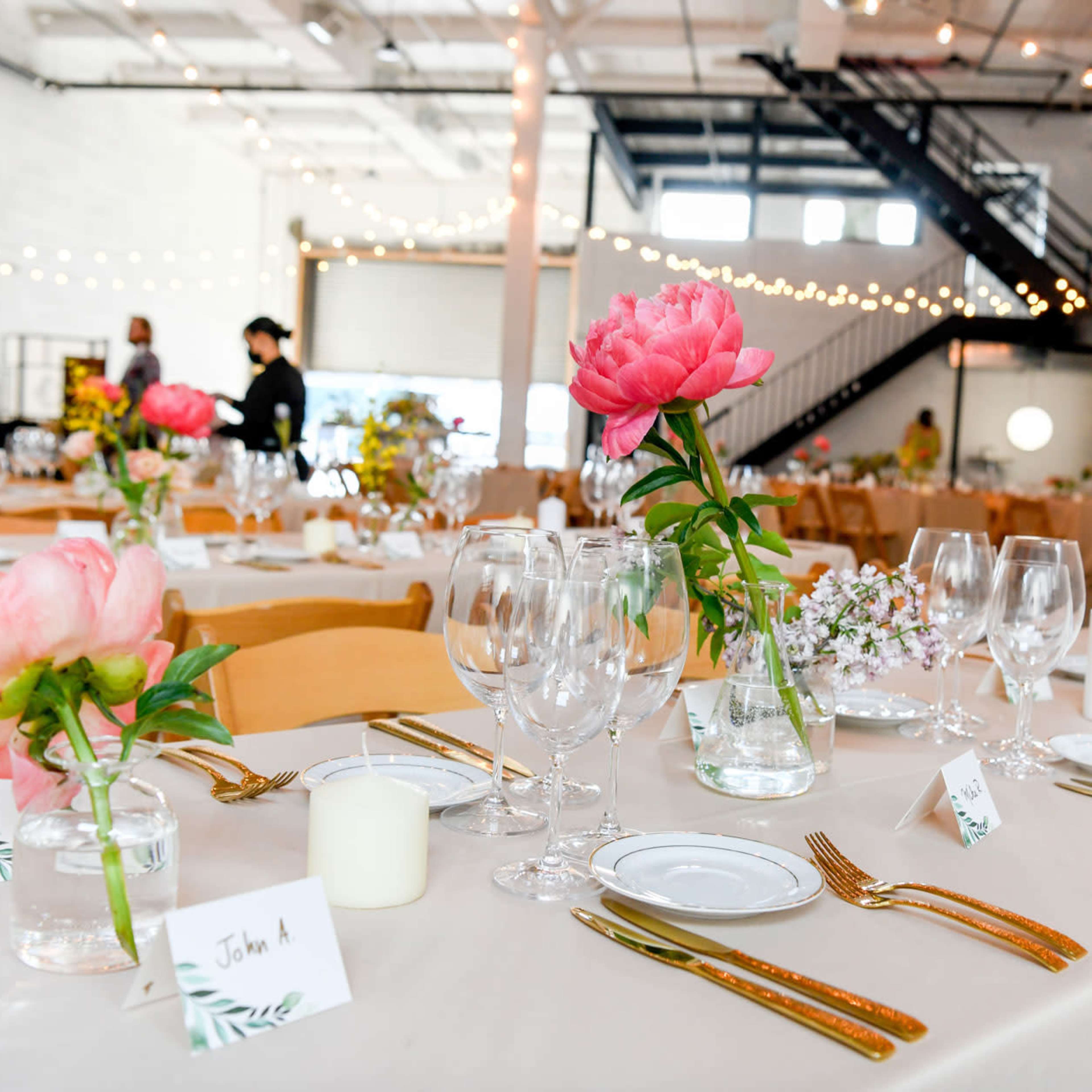 A beautifully arranged dining setup with floral centerpieces, glassware, and place cards in a spacious, decorated indoor venue.