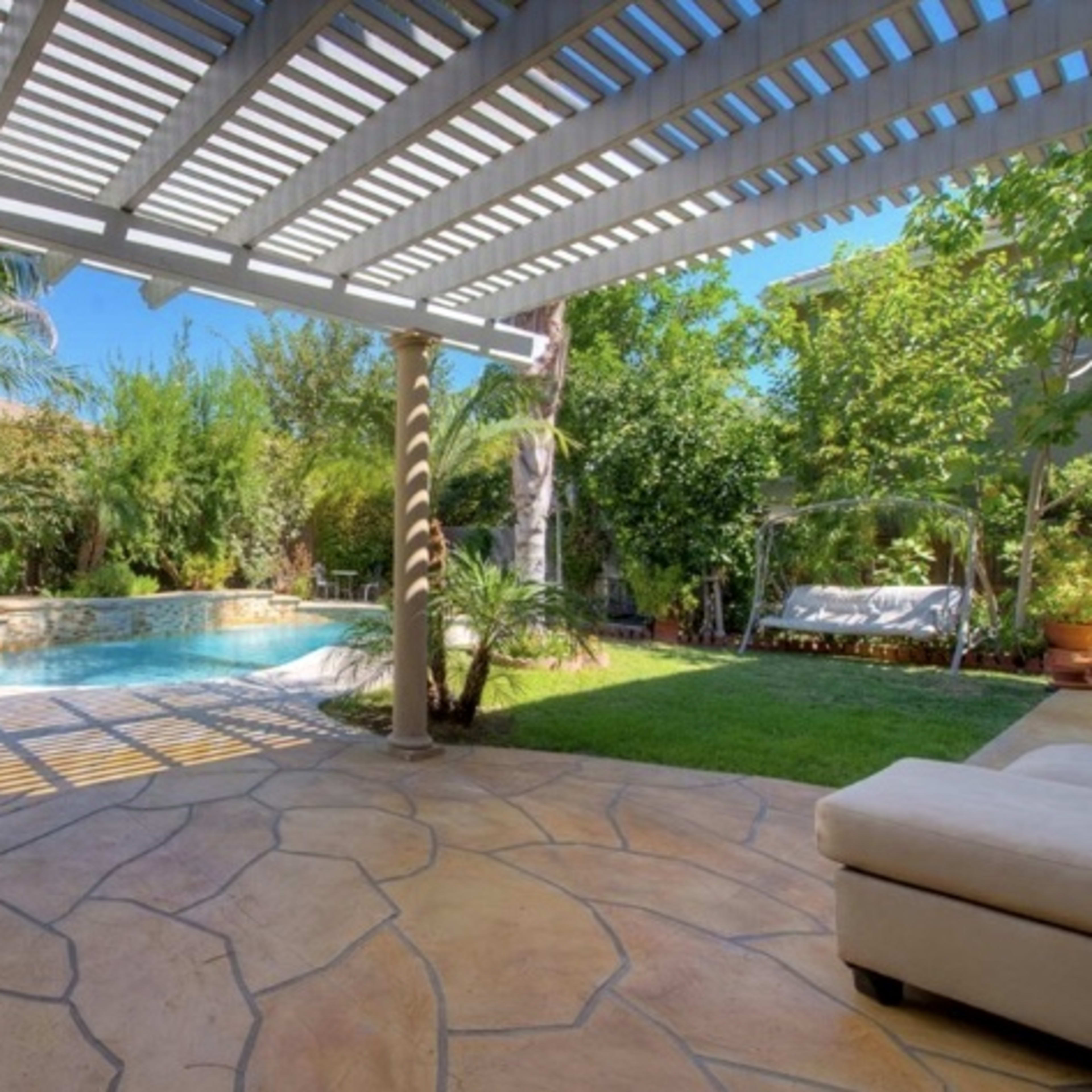 A backyard with a swimming pool surrounded by greenery and shaded by a pergola.