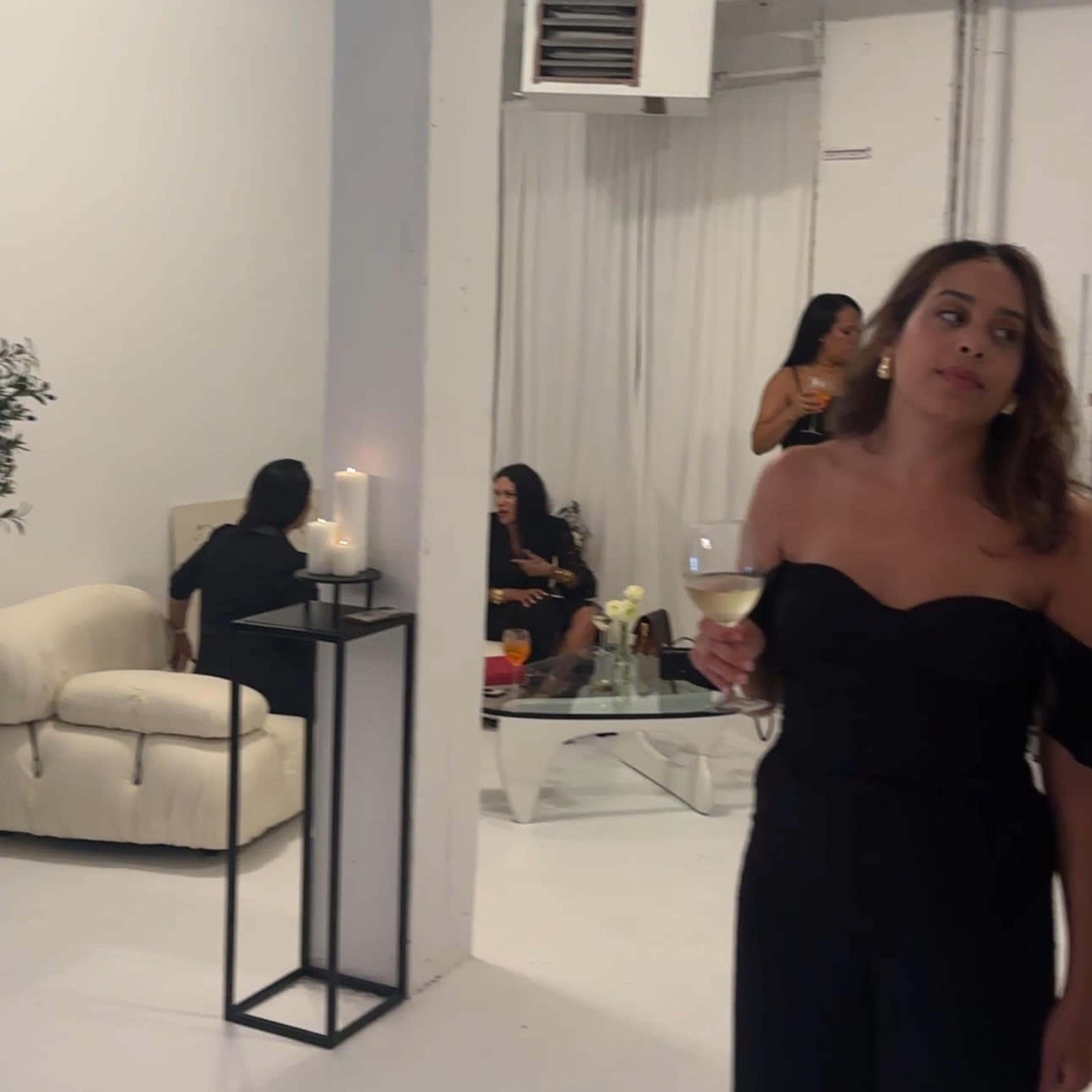 A woman in a black off-shoulder dress holds a drink while standing in a brightly lit, minimalist event space with a white sofa and guests in the background.