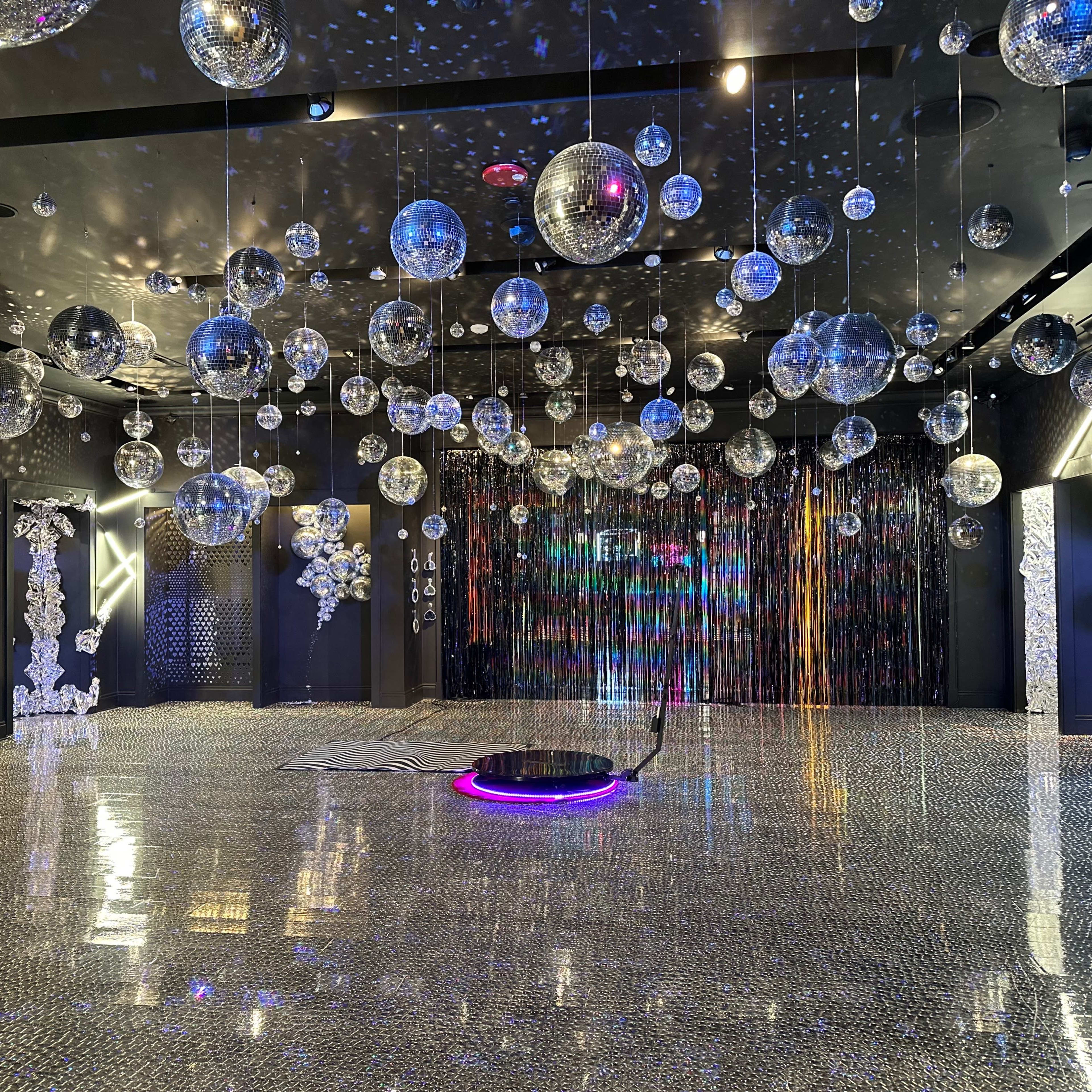 A spacious room adorned with numerous mirrored disco balls hanging from the ceiling, illuminated by colorful lights reflecting off a tiled floor.