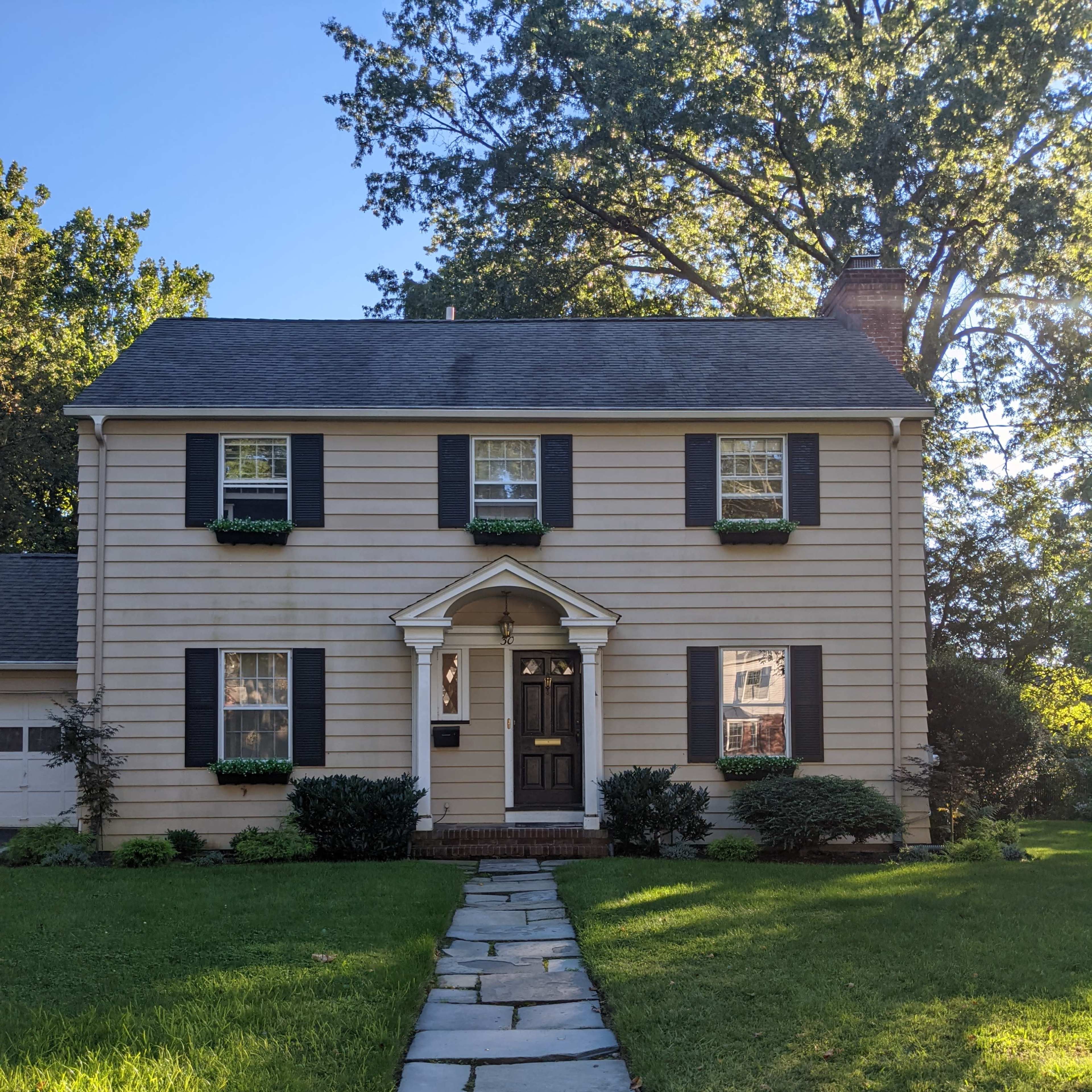 A two-story, light-colored house with black shutters and a front walkway made of stone leads to a central entrance.