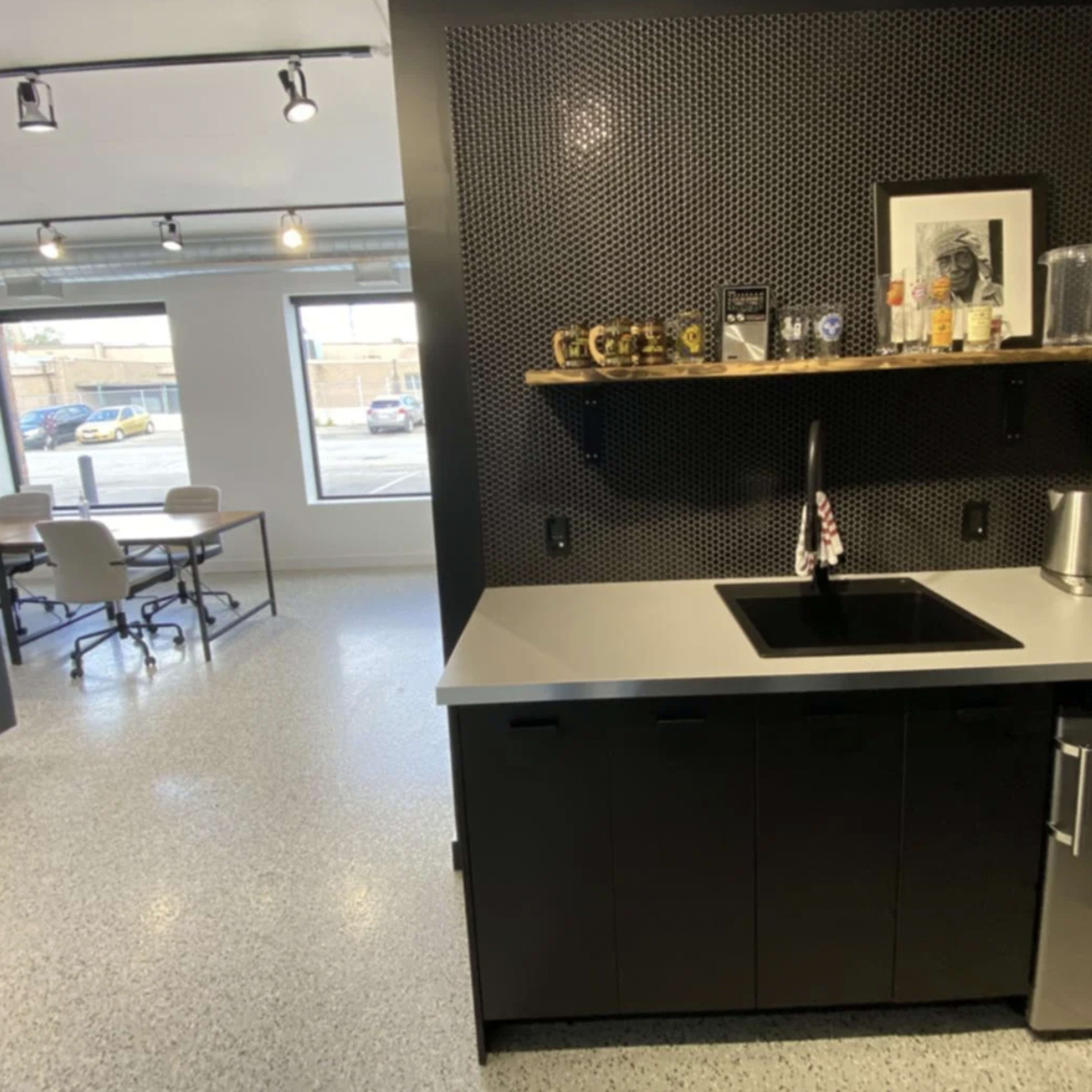 A modern kitchenette with a black countertop, a sink, a shelf displaying various items, and a small refrigerator, adjacent to a meeting area with tables and chairs.