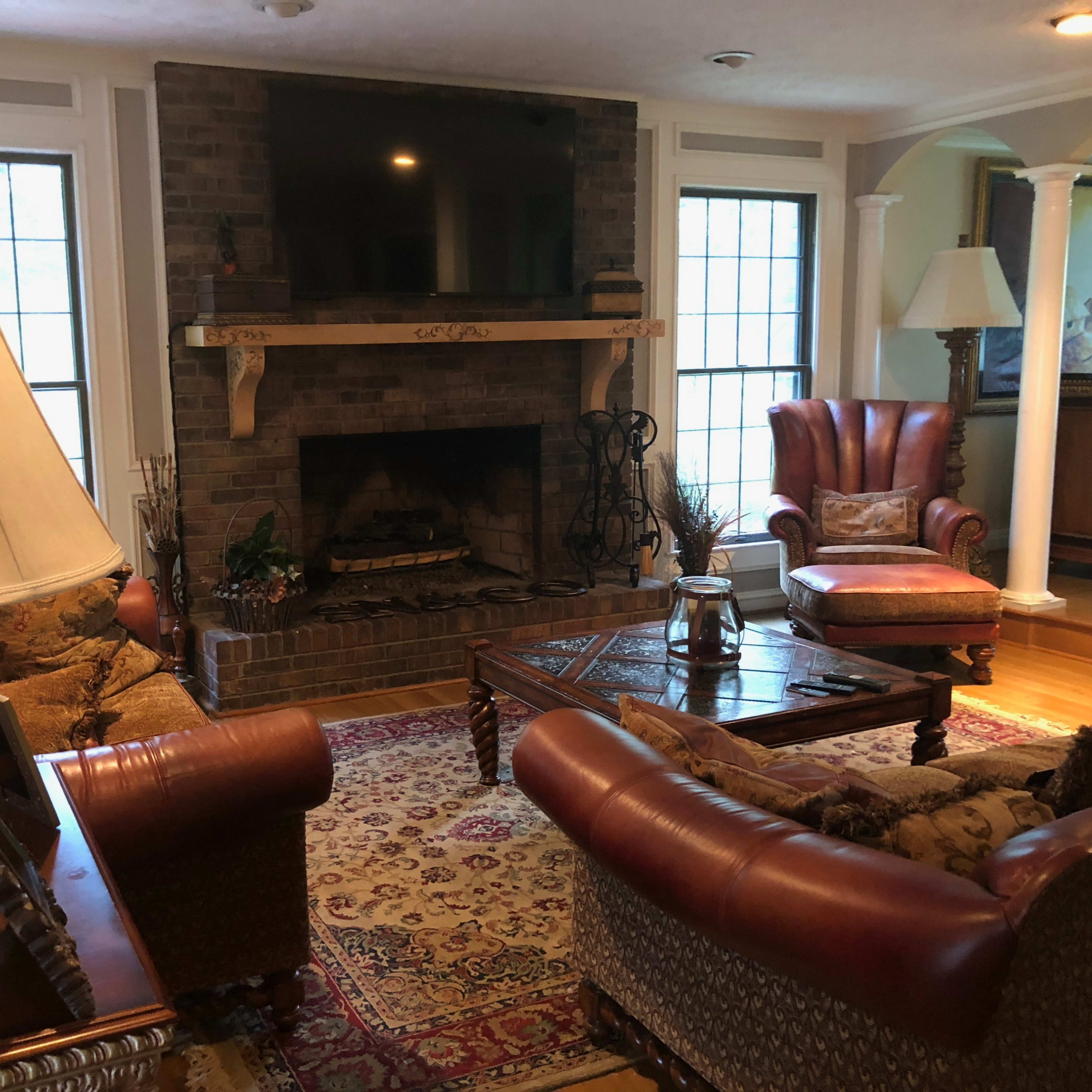A furnished living room with a stone fireplace, two armchairs, and a coffee table on a patterned rug, illuminated by natural light from large windows.