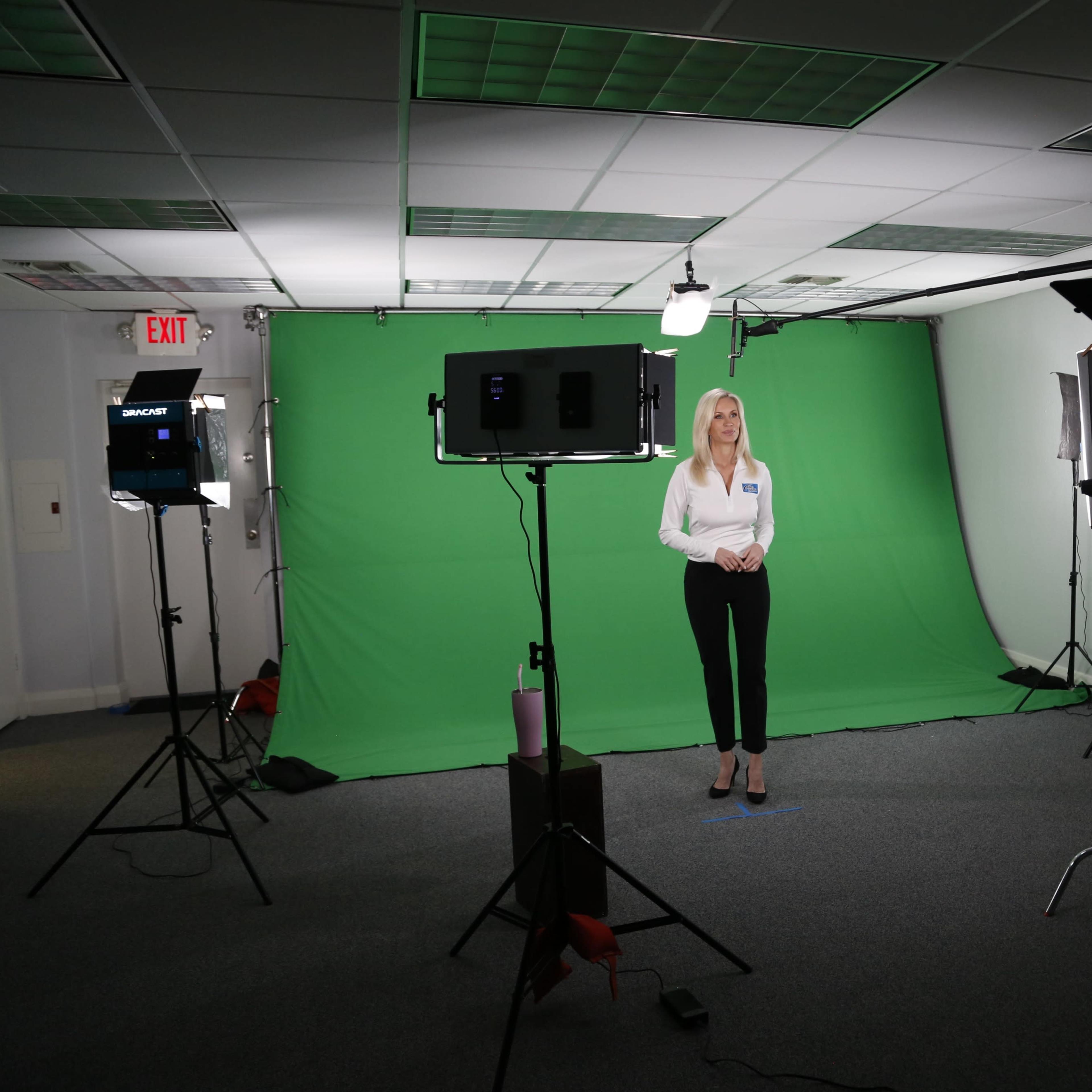 A woman stands in front of a green screen in a studio, surrounded by cameras and lighting equipment.