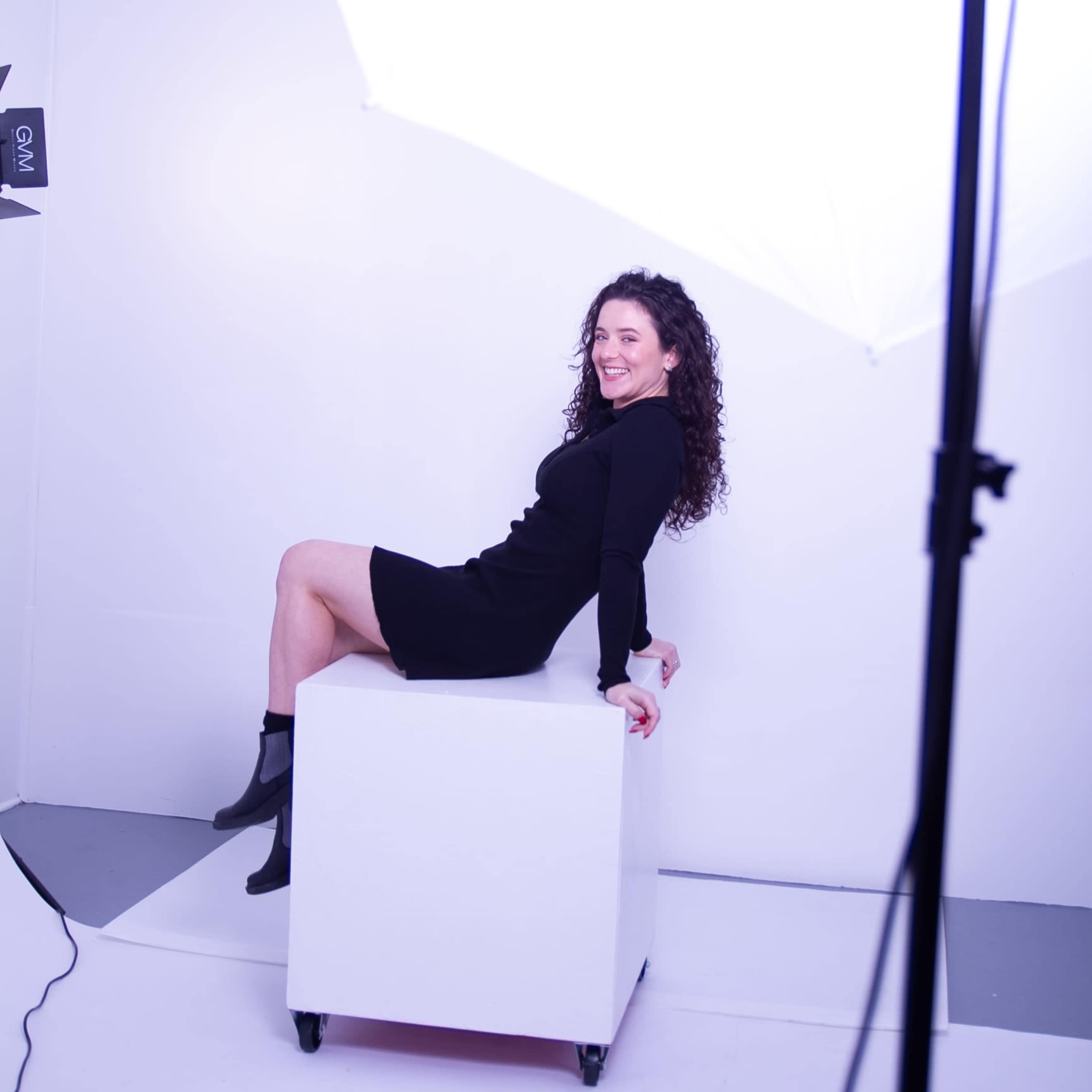 A woman sits on a white cube in a photography studio, with bright lights illuminating the scene.