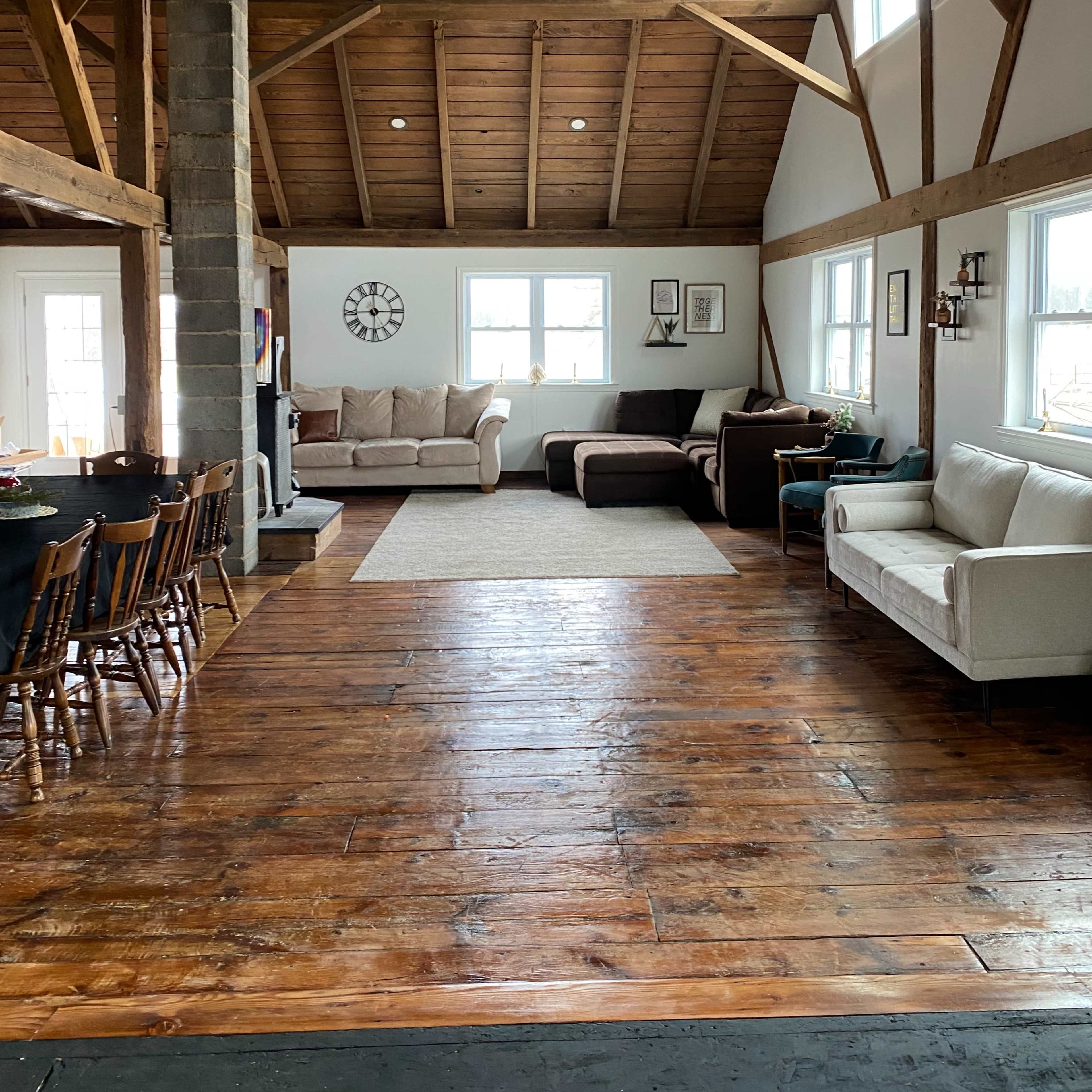 A spacious, well-lit living area with wooden beams, a rustic wood floor, multiple sofas, and a dining table with chairs.