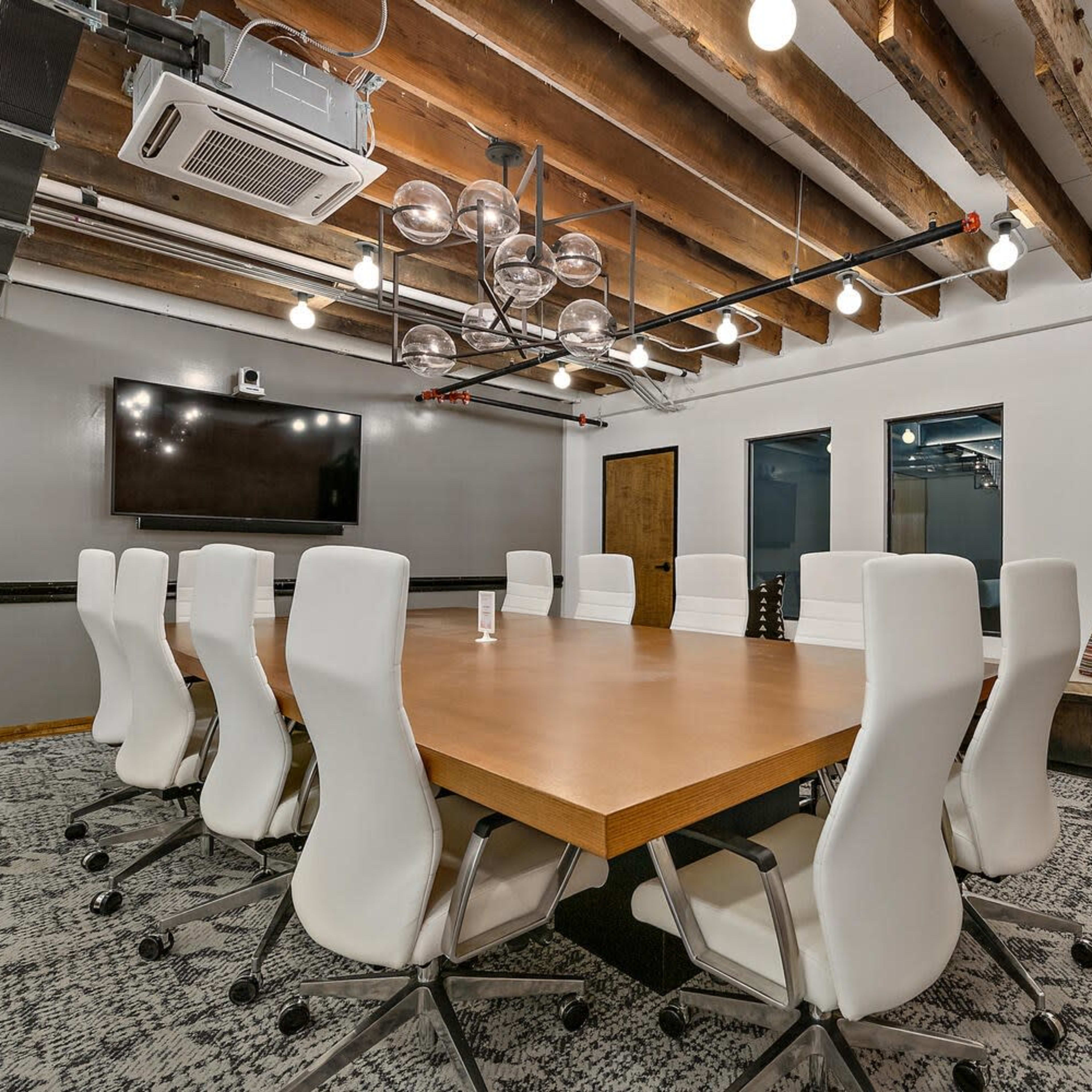 A conference room features a large wooden table surrounded by white chairs, with modern lighting and a television on the wall.