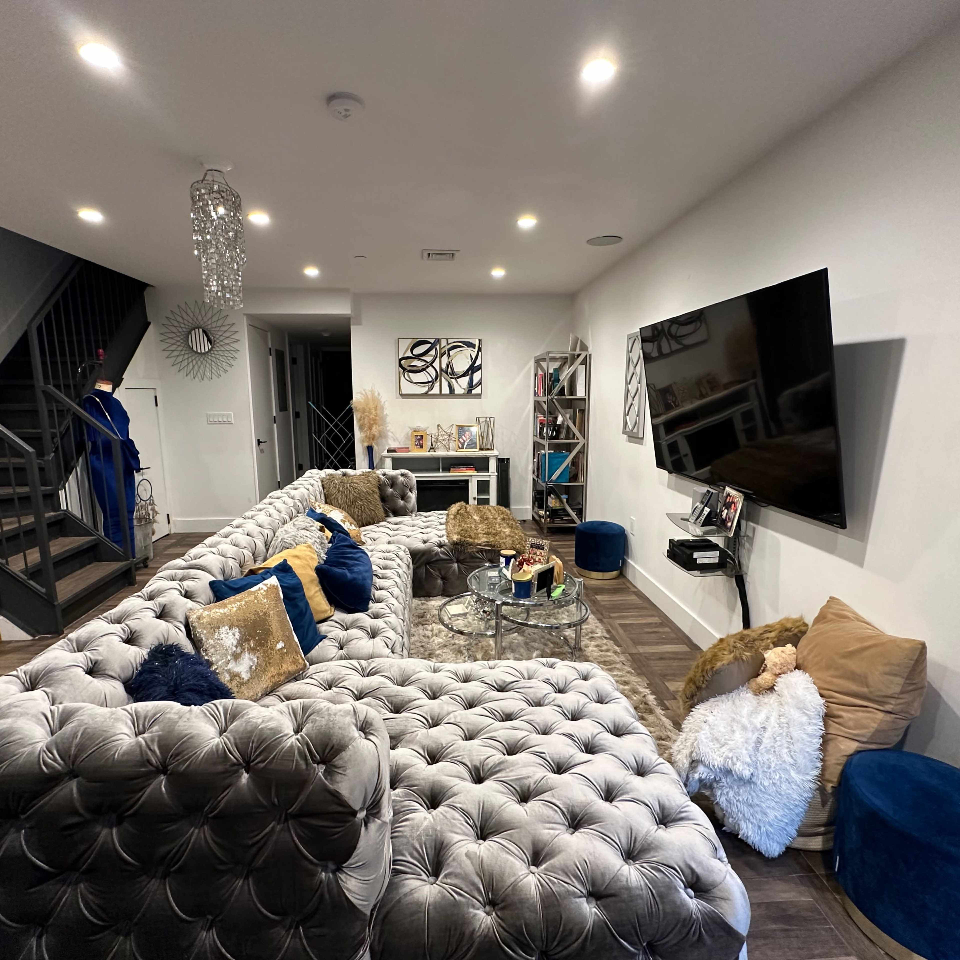 A modern living room features a large tufted sectional sofa, a coffee table, a wall-mounted television, and decorative elements along with a staircase in the background.