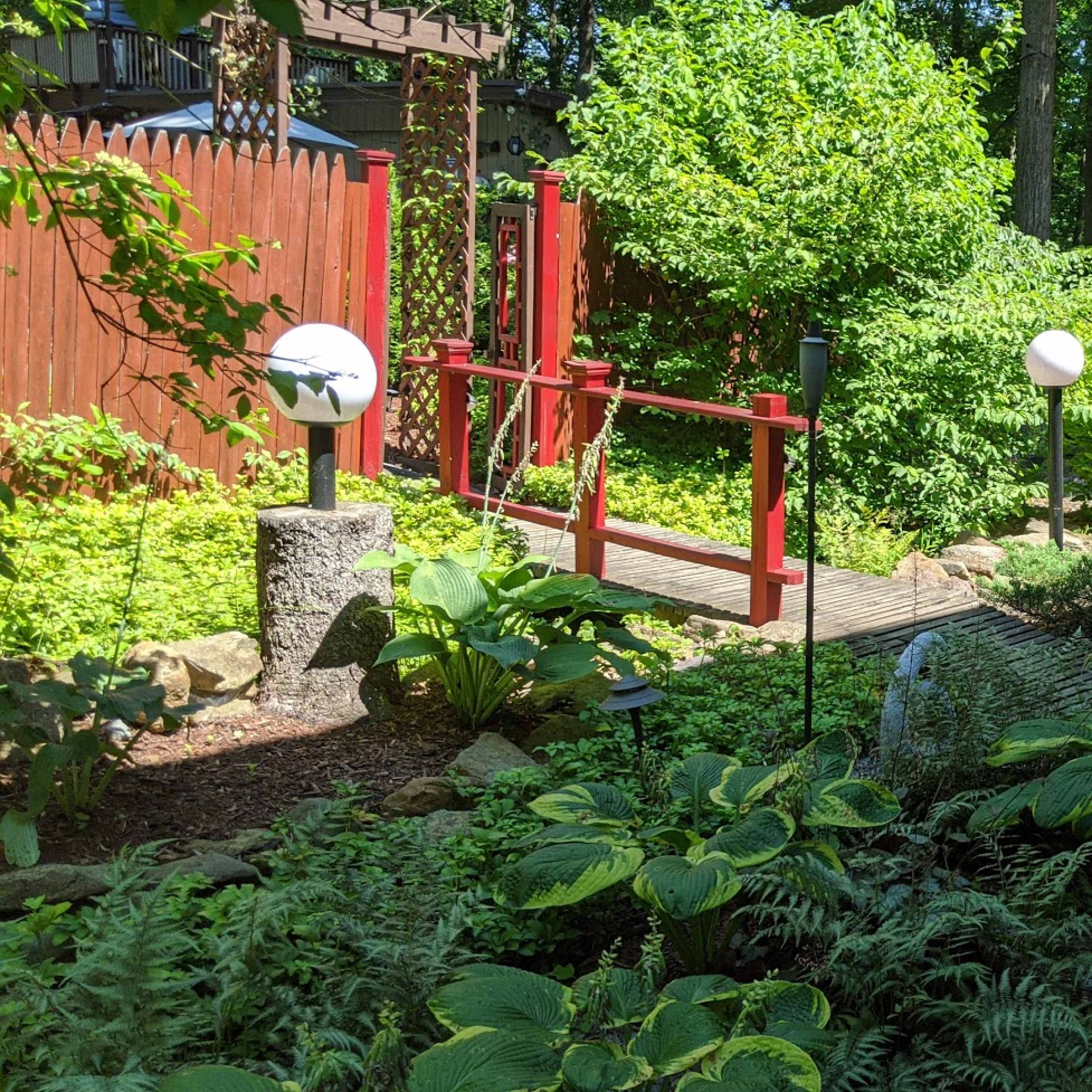 A landscaped garden with a wooden bridge, various plants, and decorative lighting elements amidst trees.