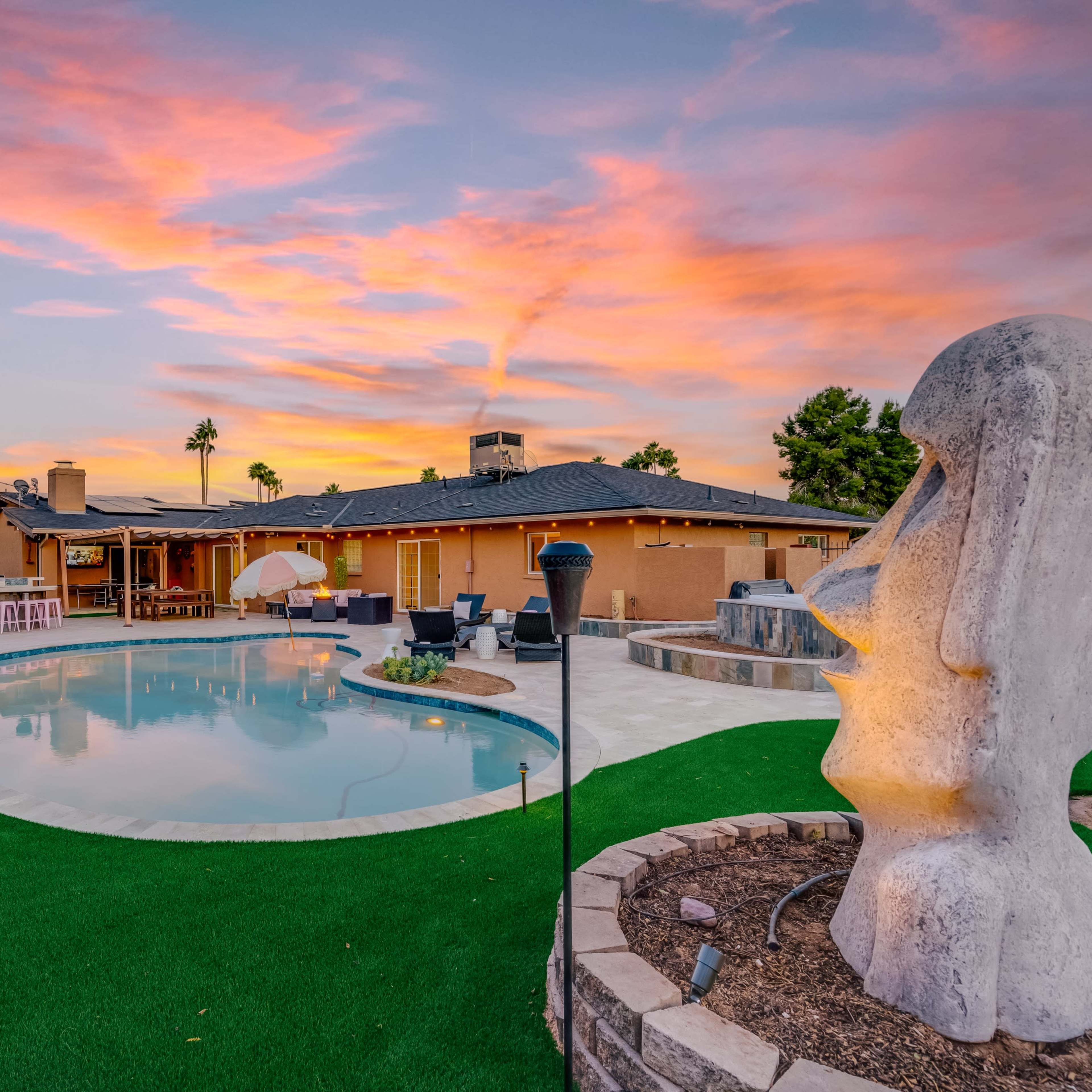 A modern backyard with a swimming pool, landscaping, lounge chairs, and a large decorative sculpture, set against a colorful sunset sky.