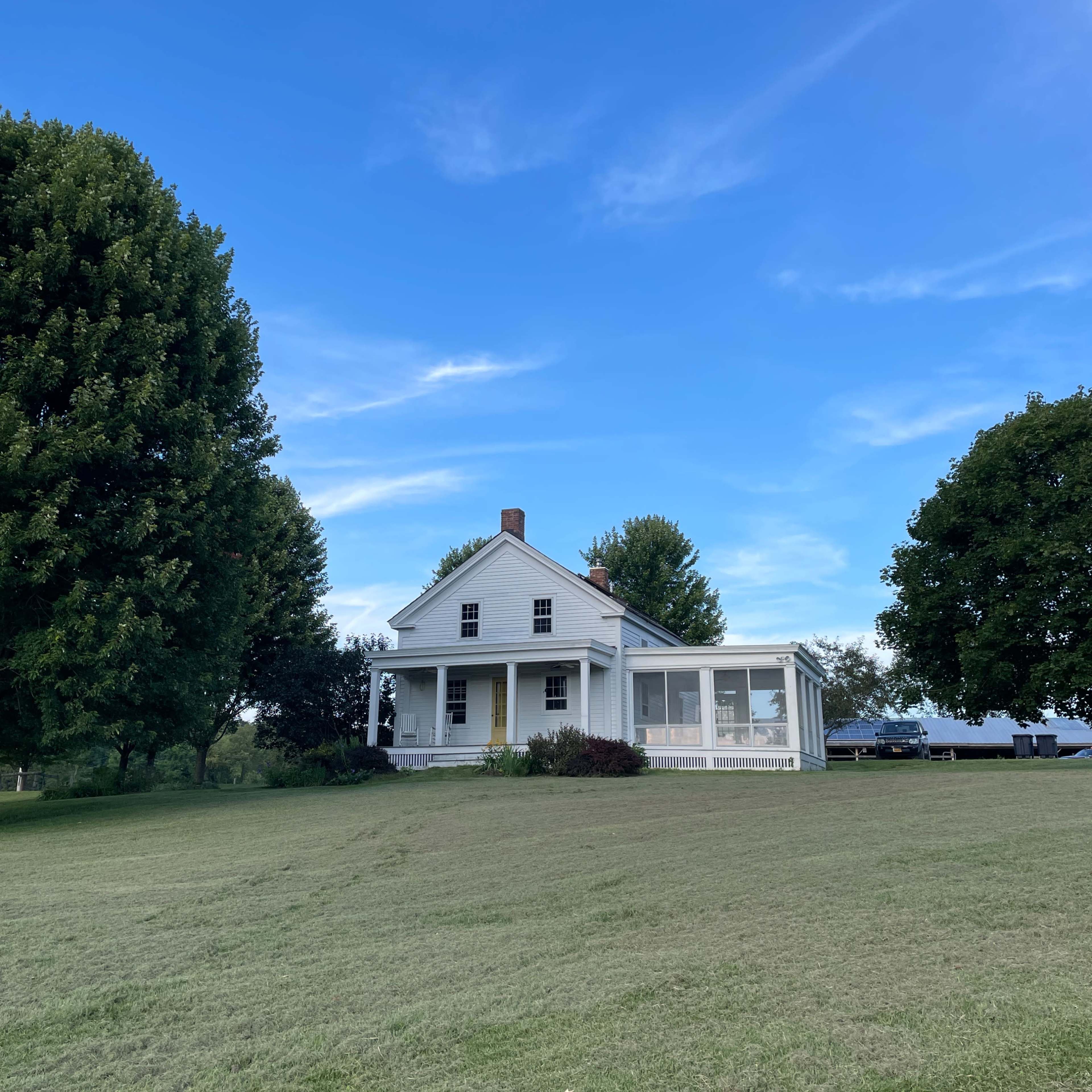 A white two-story house with a porch is surrounded by green grass and trees under a blue sky.