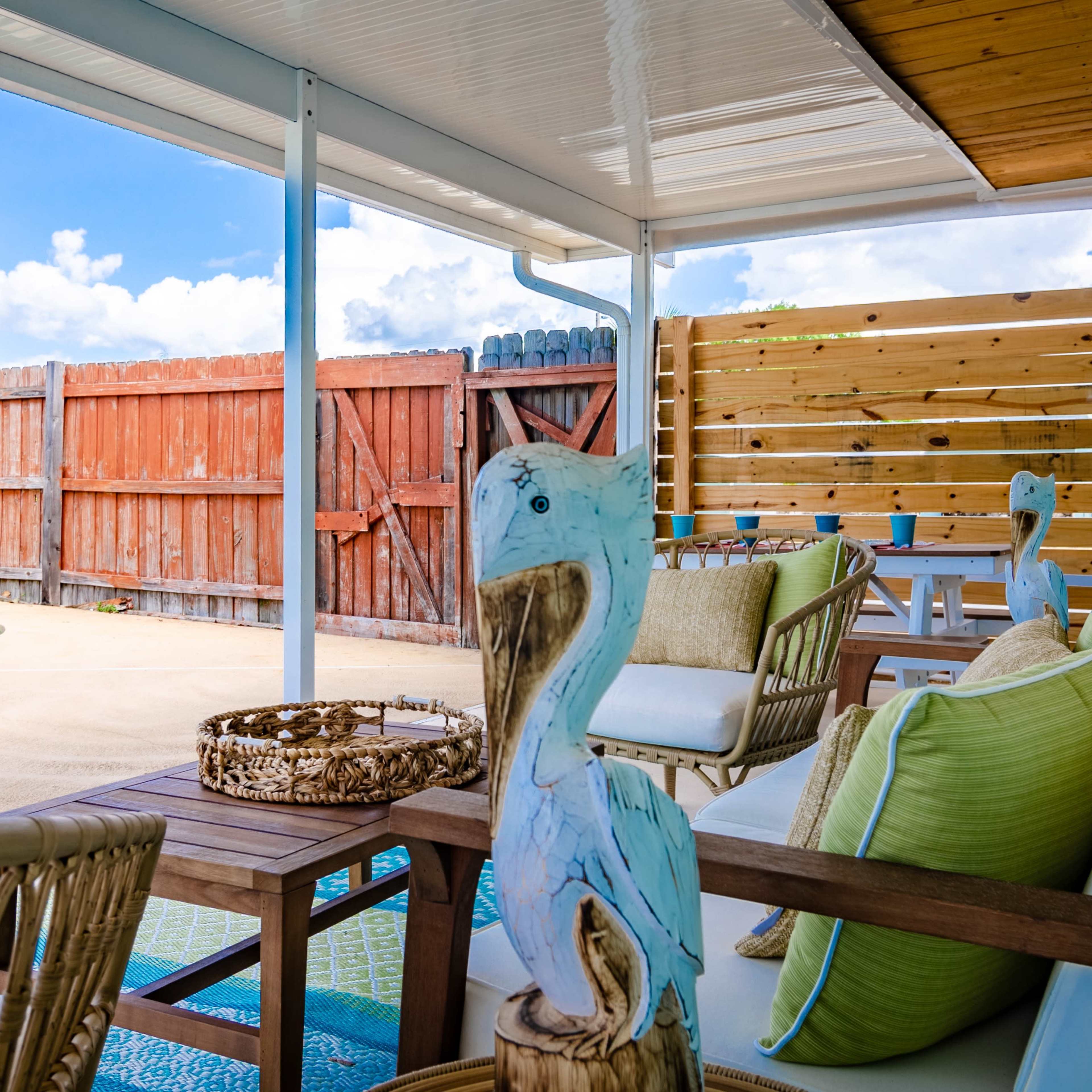 A shaded patio with wicker chairs and decorative pelican figures overlooks a swimming pool surrounded by a wooden fence.