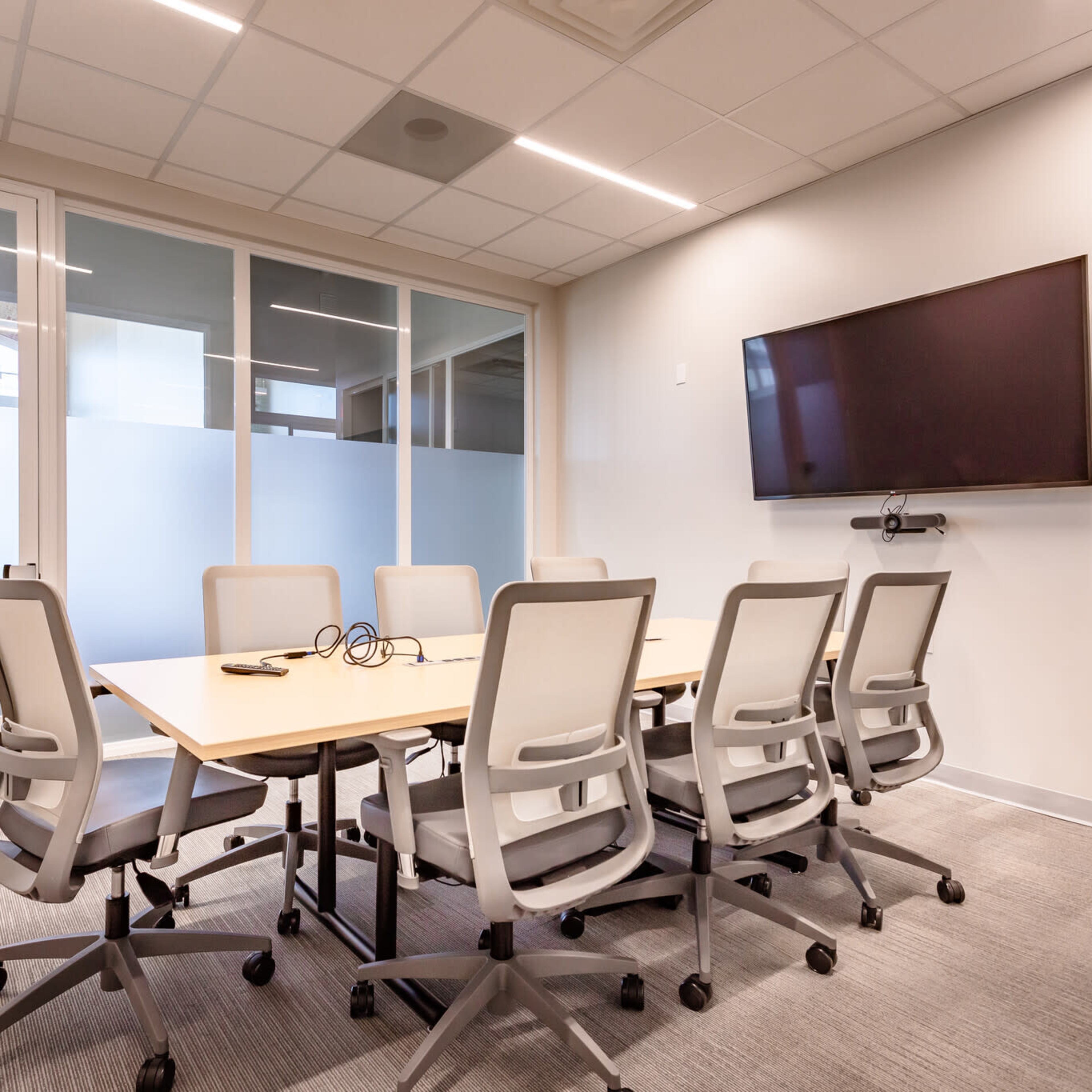 A modern conference room features a long table surrounded by six ergonomic chairs, with a wall-mounted TV and glass-paneled doors.