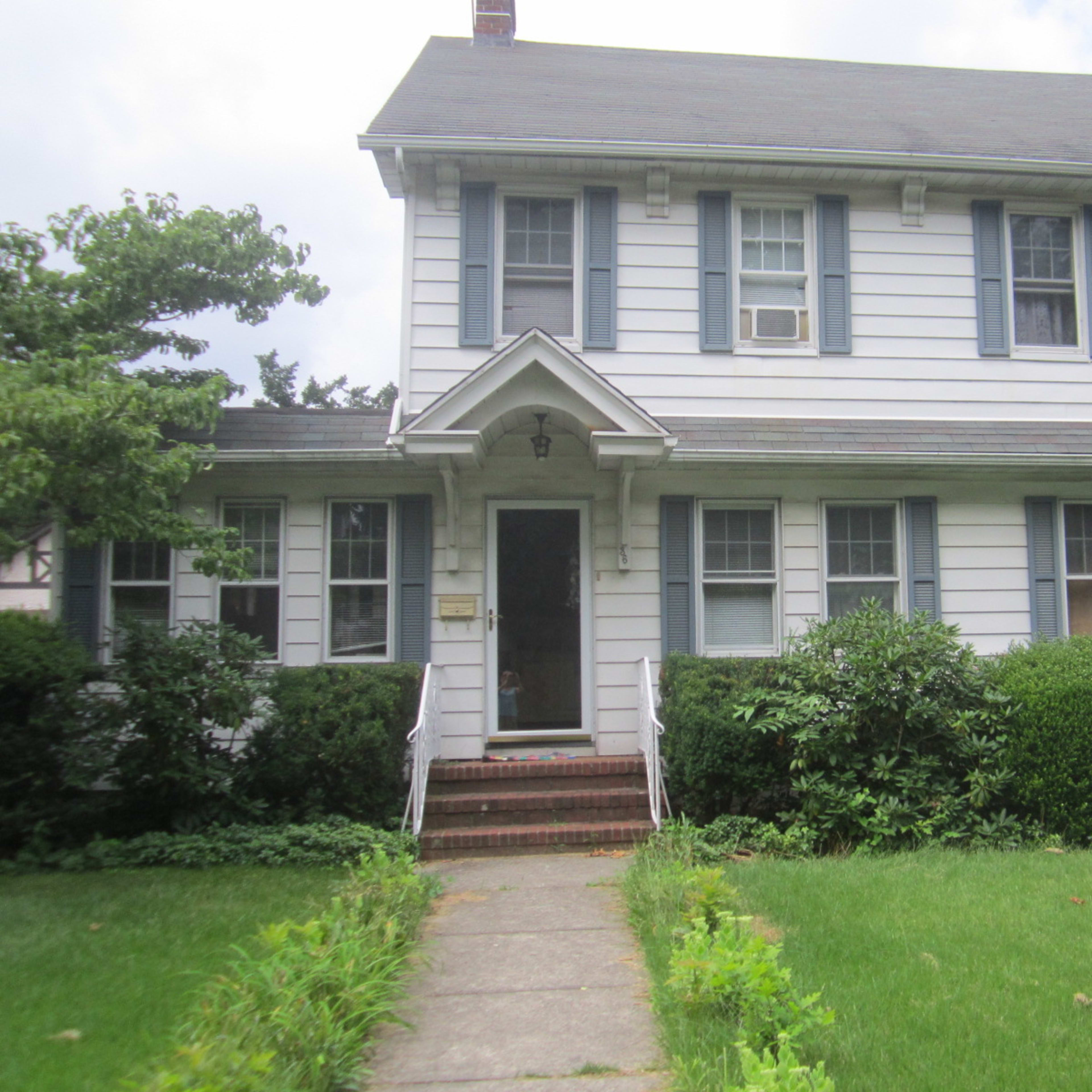 A two-story, white wooden house with dark blue shutters and a small front porch surrounded by green shrubs and a lawn.