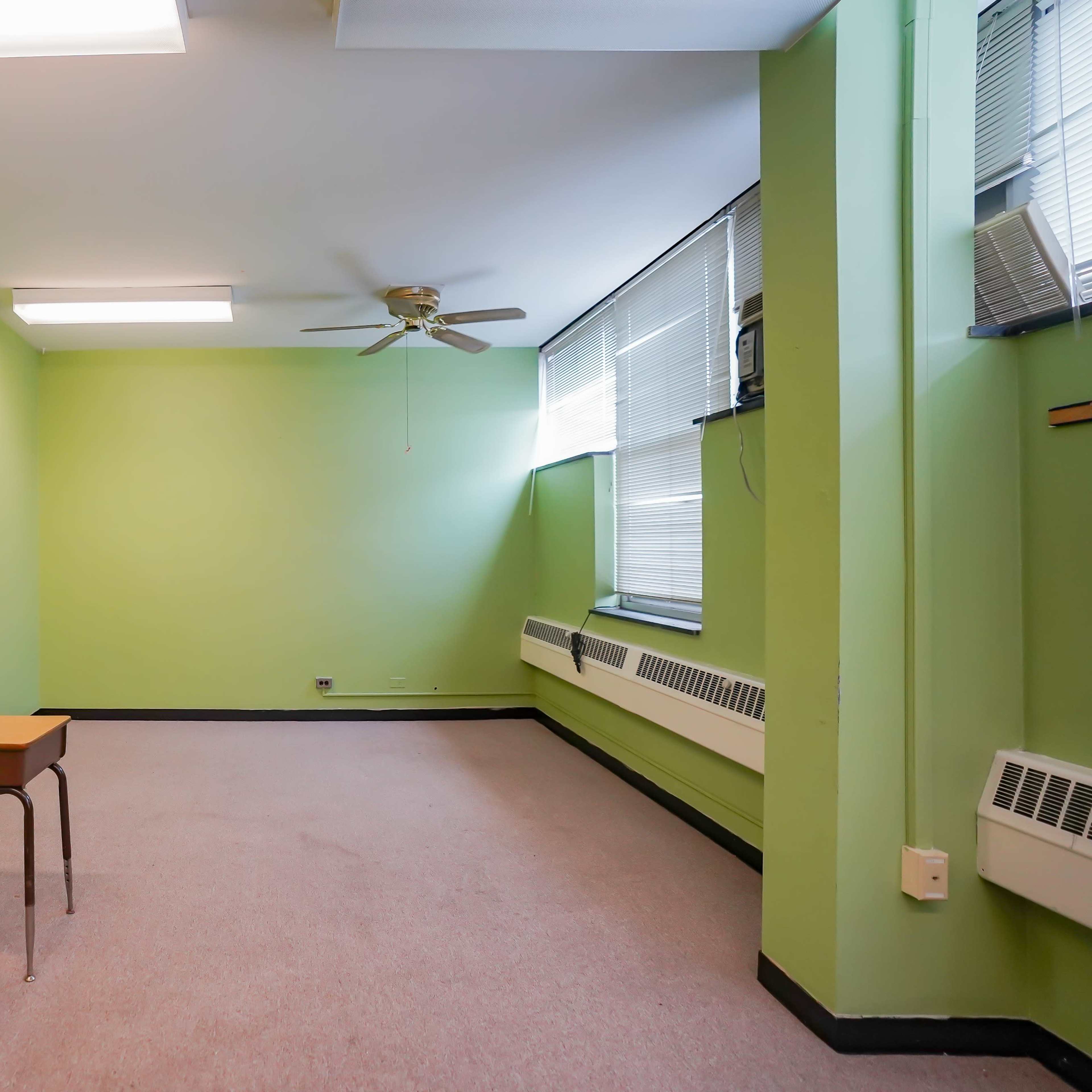 An empty room with green walls, a ceiling fan, and a single wooden desk in the center.