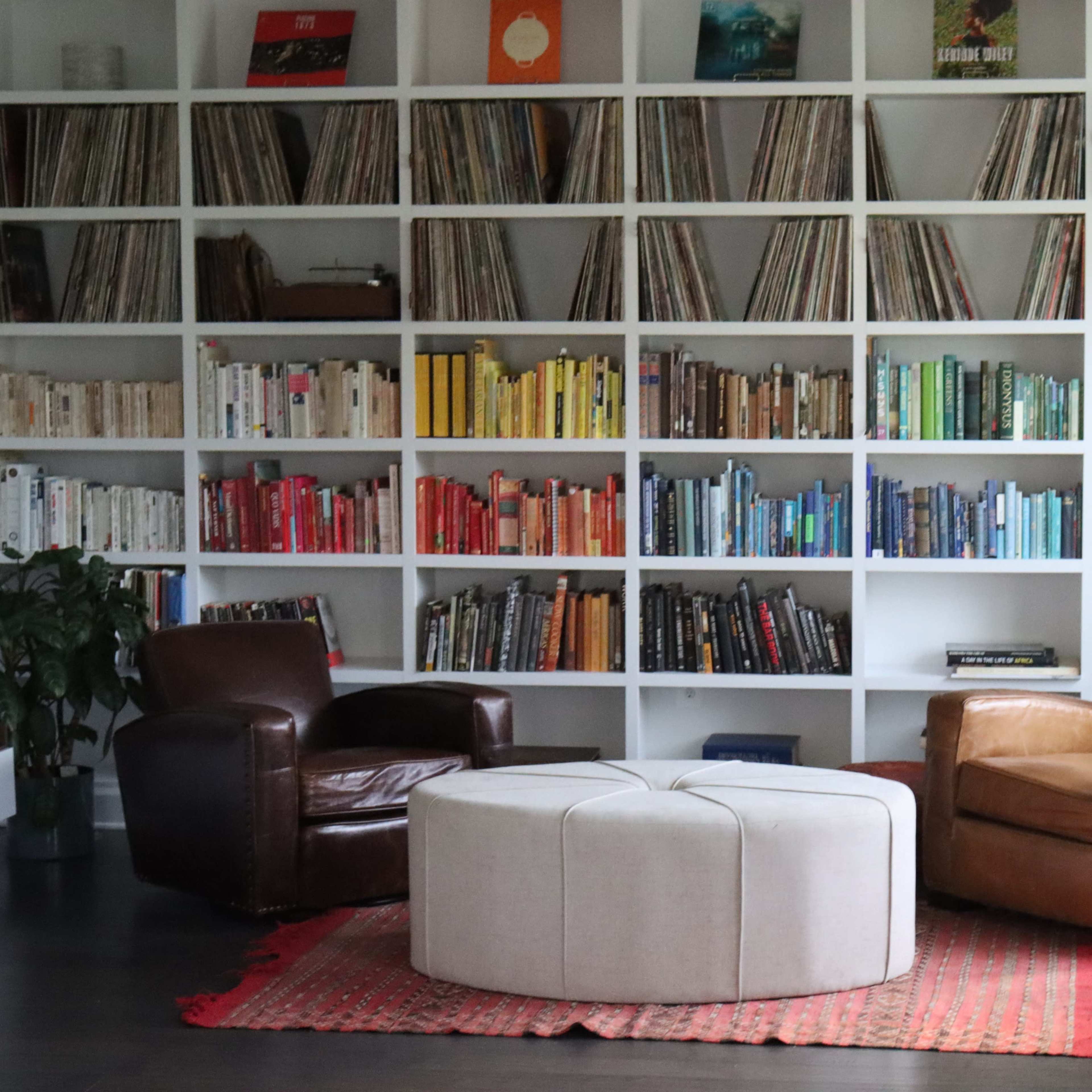 A cozy reading nook features two leather armchairs facing a circular ottoman, surrounded by a wall of books organized by color.