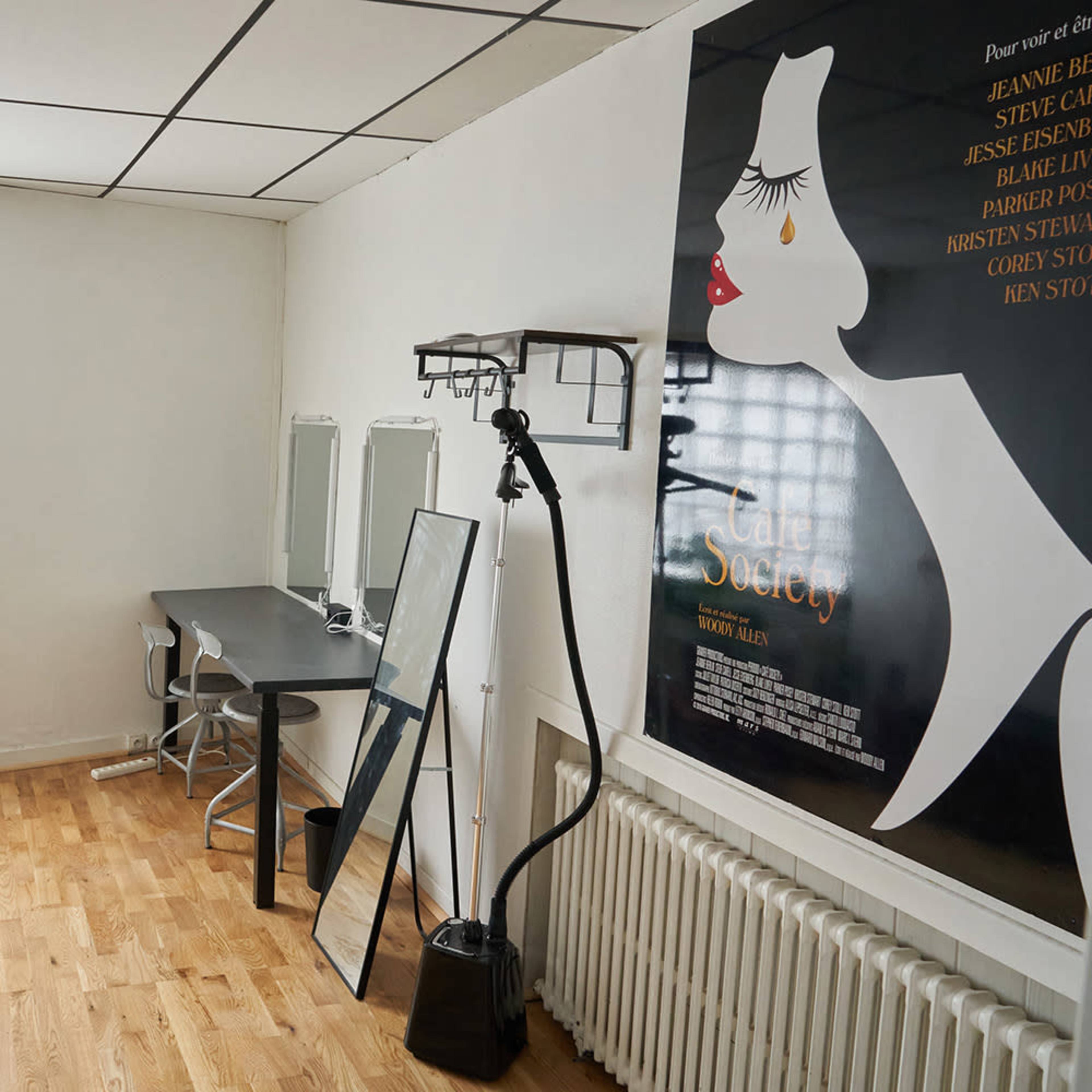 A bright room featuring a desk with a chair, a large mirror, a potted plant, and a film poster on the wall.