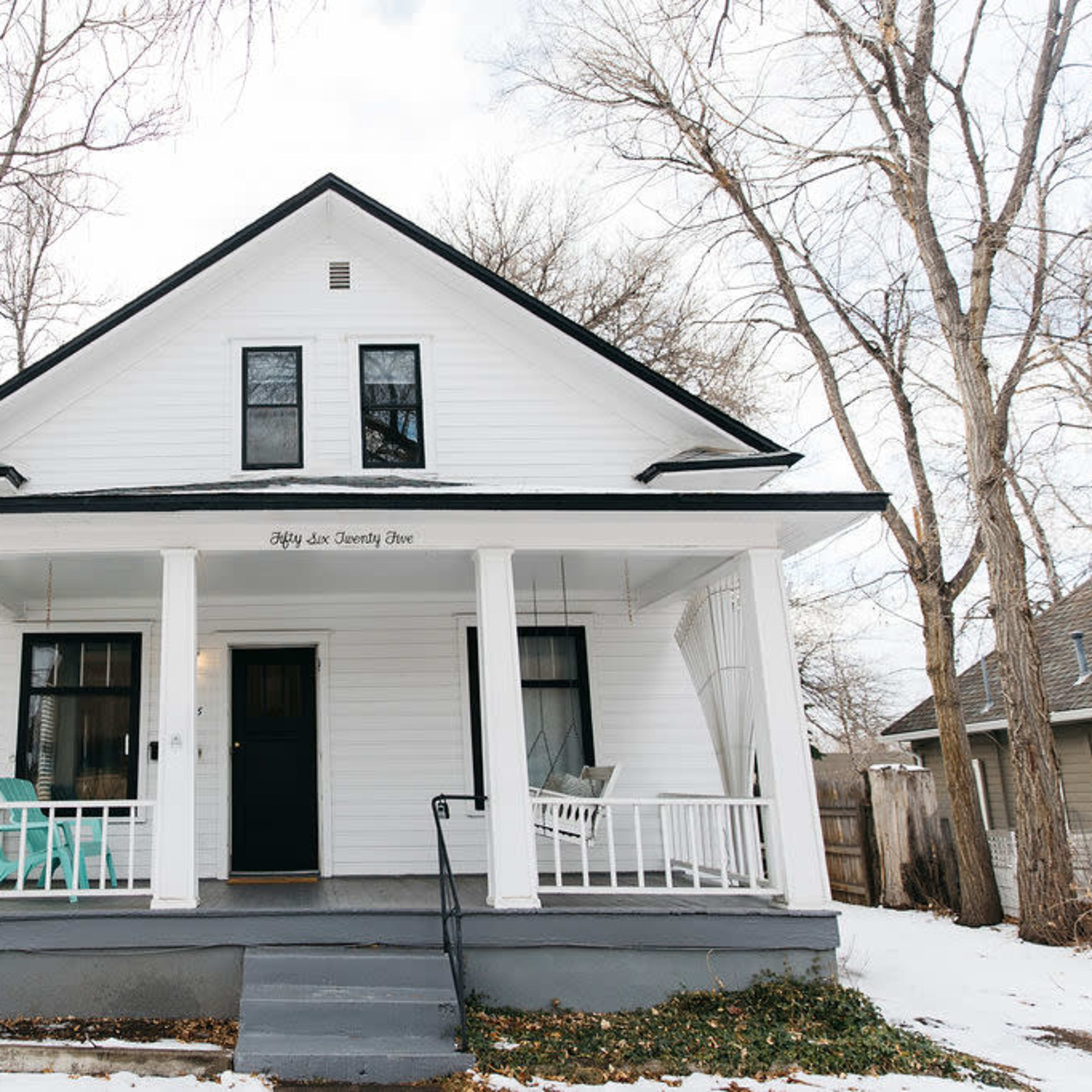 A white two-story house with a covered porch and two turquoise chairs sits among leafless trees on a quiet street.