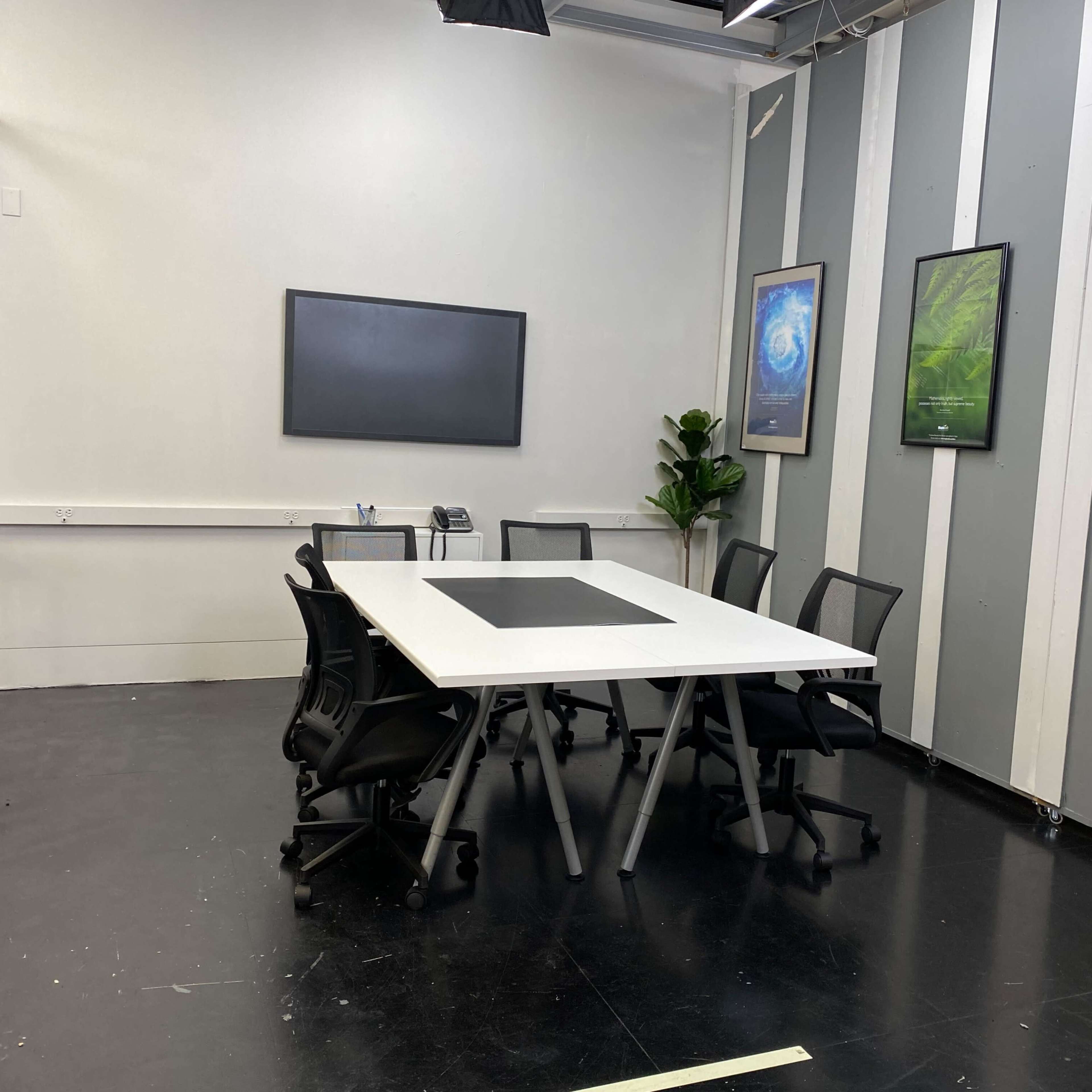 A modern conference room with a large white table surrounded by black office chairs, a wall-mounted screen, and framed nature posters.