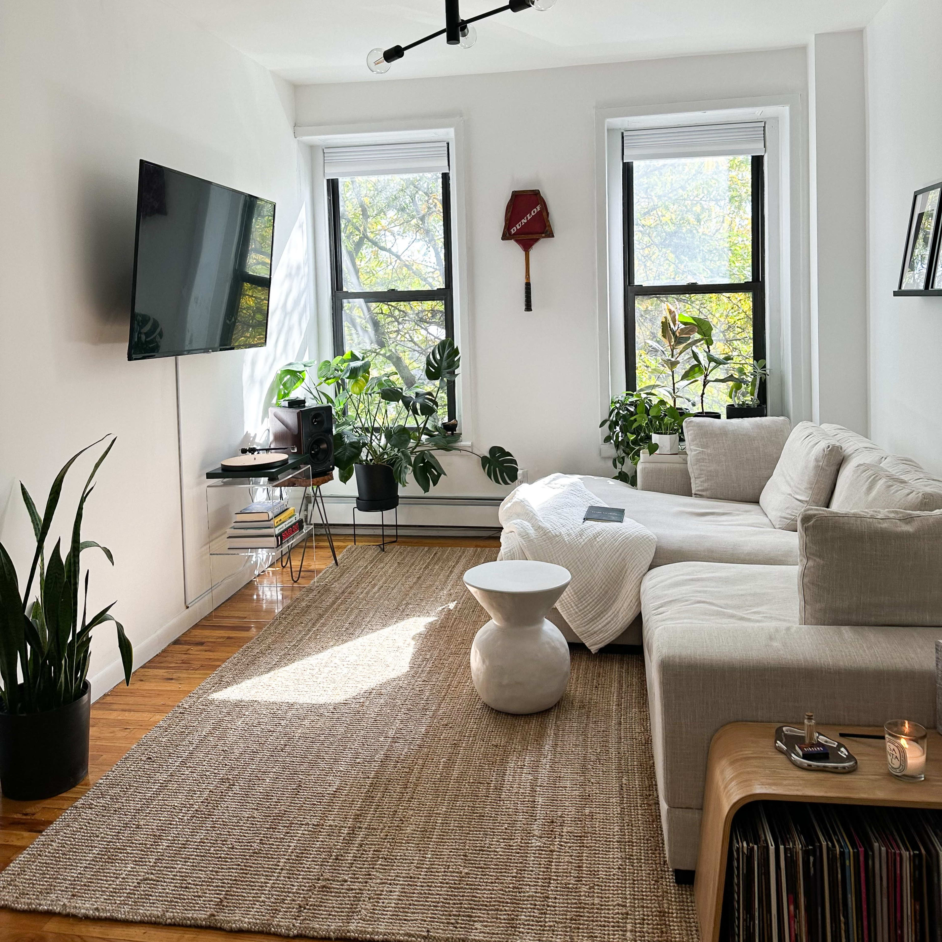 A bright living room features a sectional sofa, a television on the wall, large windows with plants, and a round coffee table.