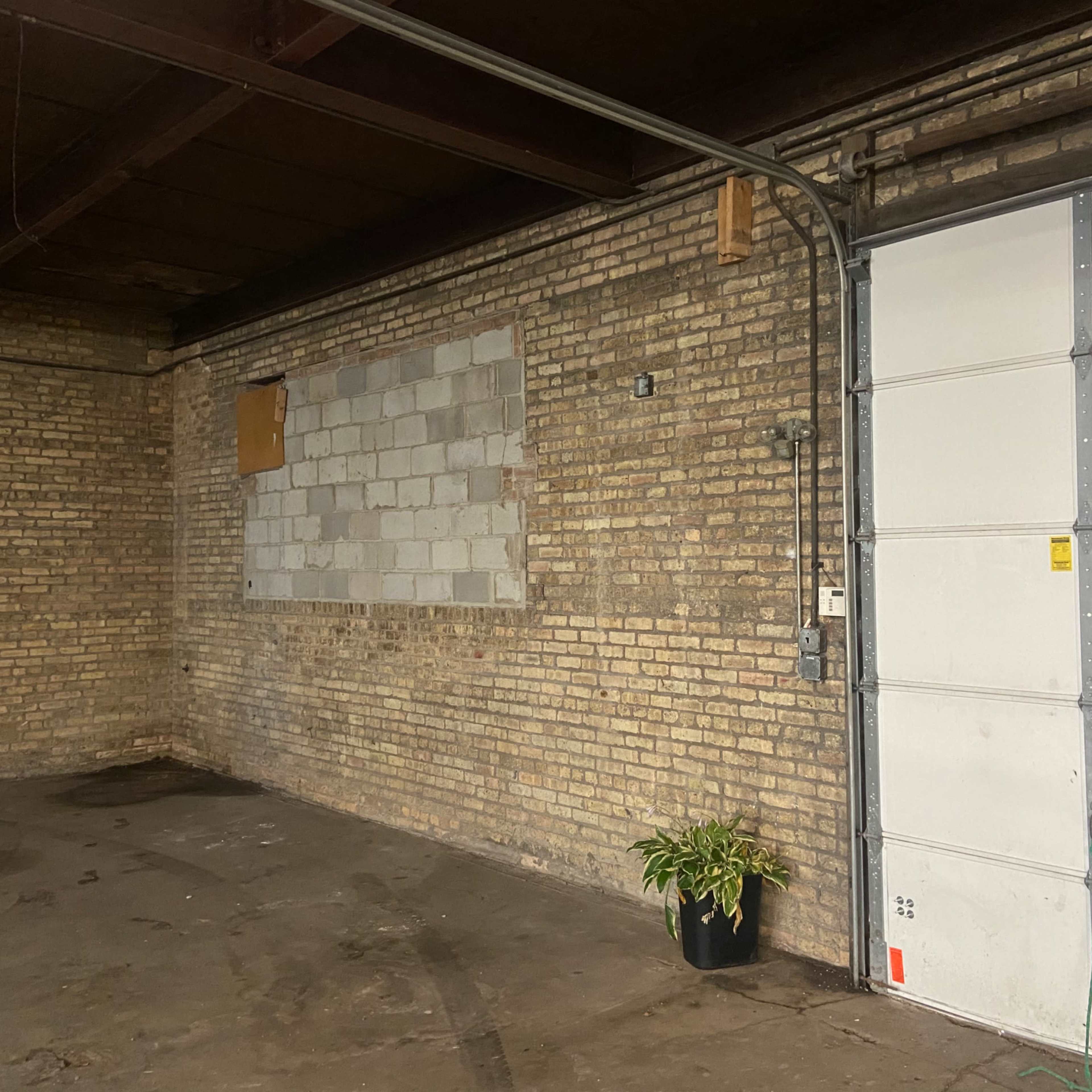A brick-walled garage features an unfinished section of wall, a roll-up door, and a potted plant in one corner.