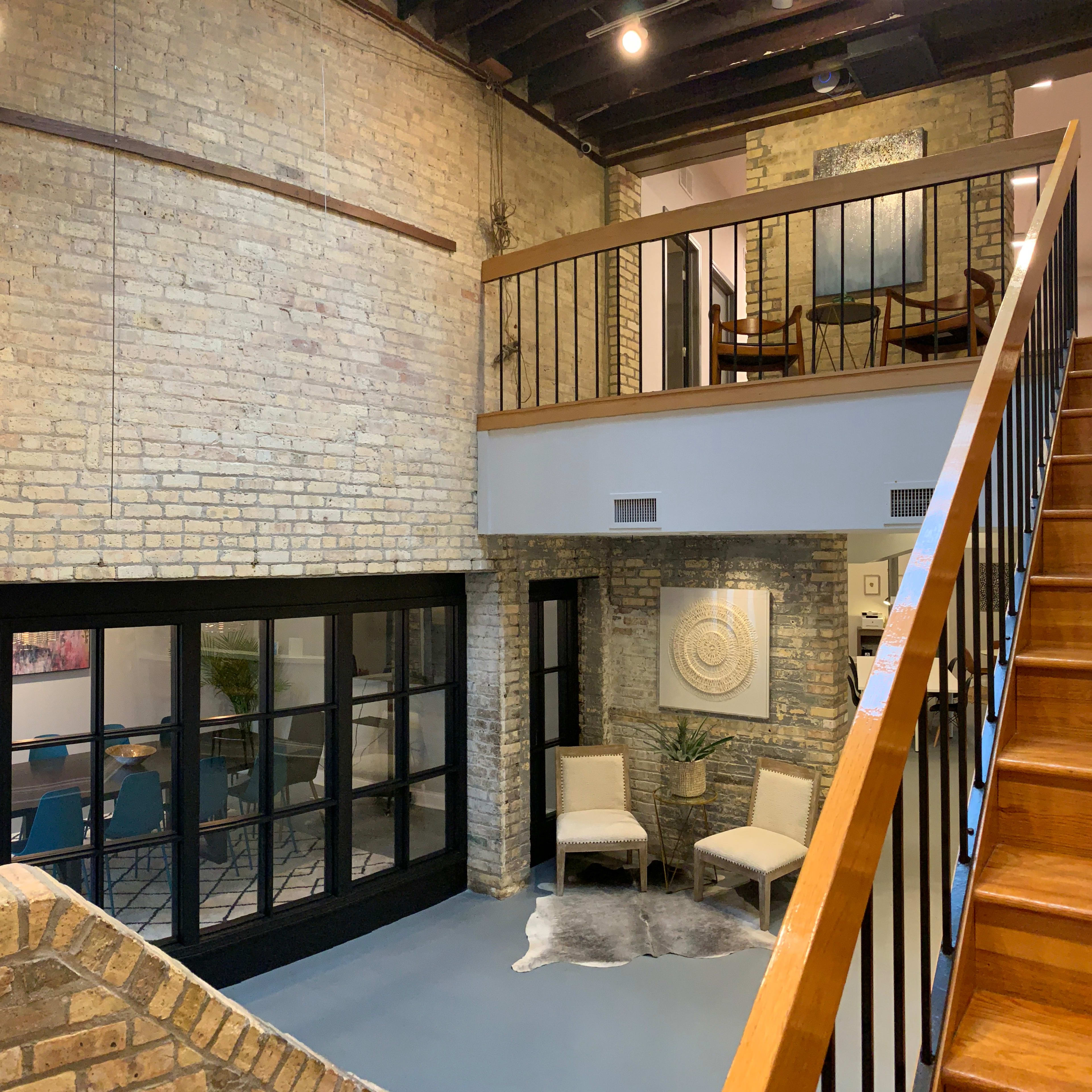 A modern interior space with exposed brick walls, a wooden staircase leading to a balcony, and a glass door opening to a room with a dining table.
