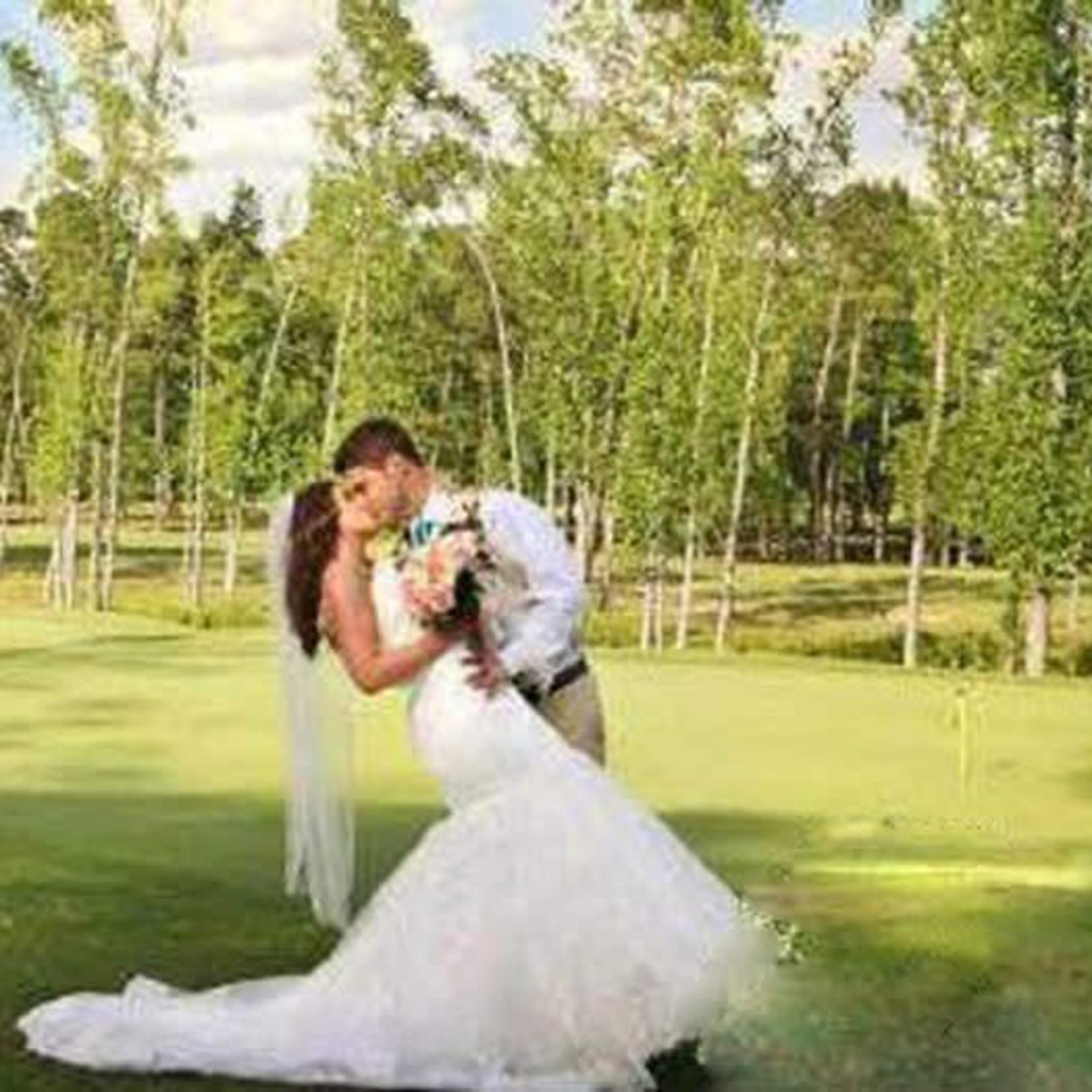 A newlywed couple embraces on a grassy area surrounded by trees.