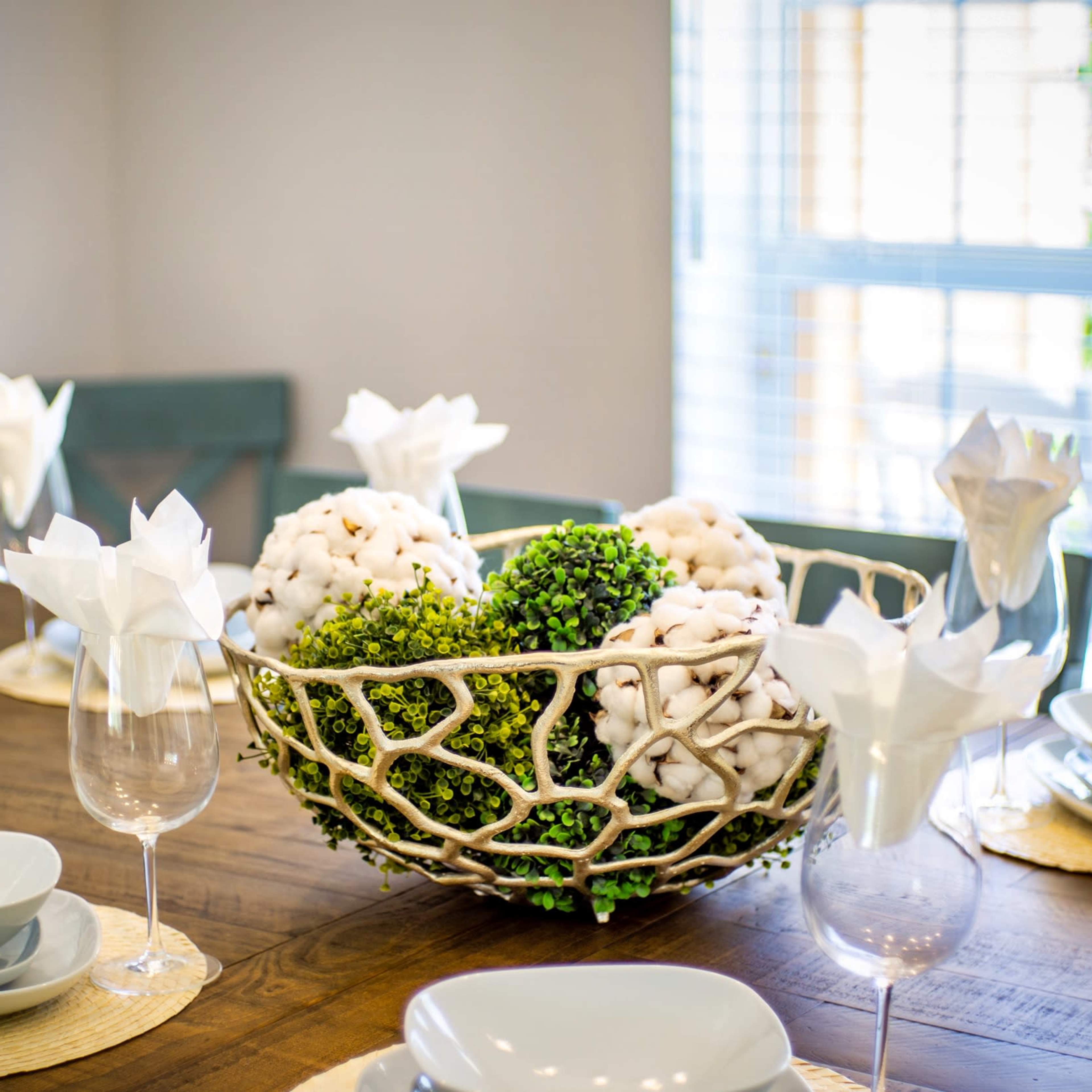 A dining table is set with grey plates and glasses, featuring a decorative bowl of cotton and greenery at the center.