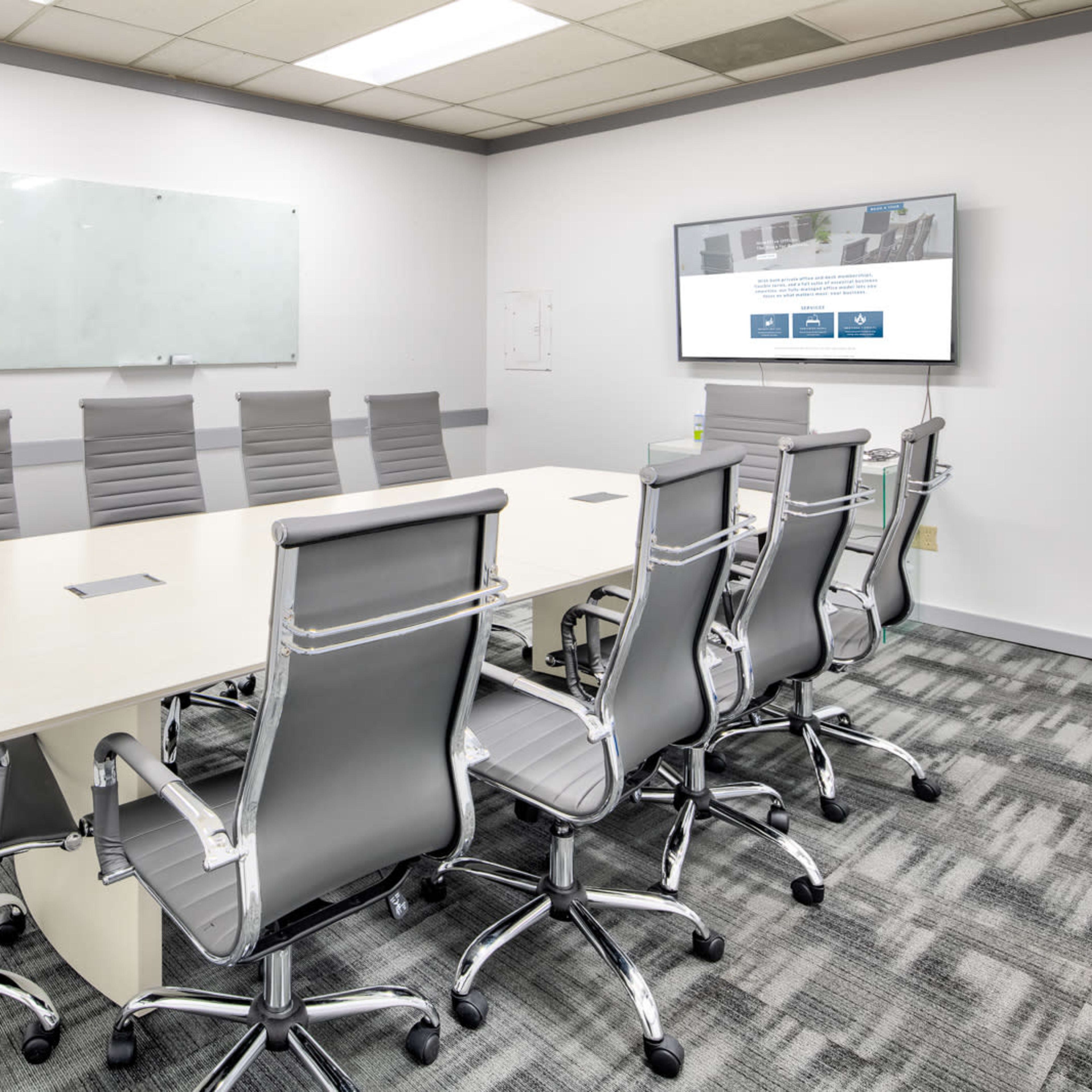 A modern conference room features a long table surrounded by ergonomic chairs, with a wall-mounted screen and a whiteboard.