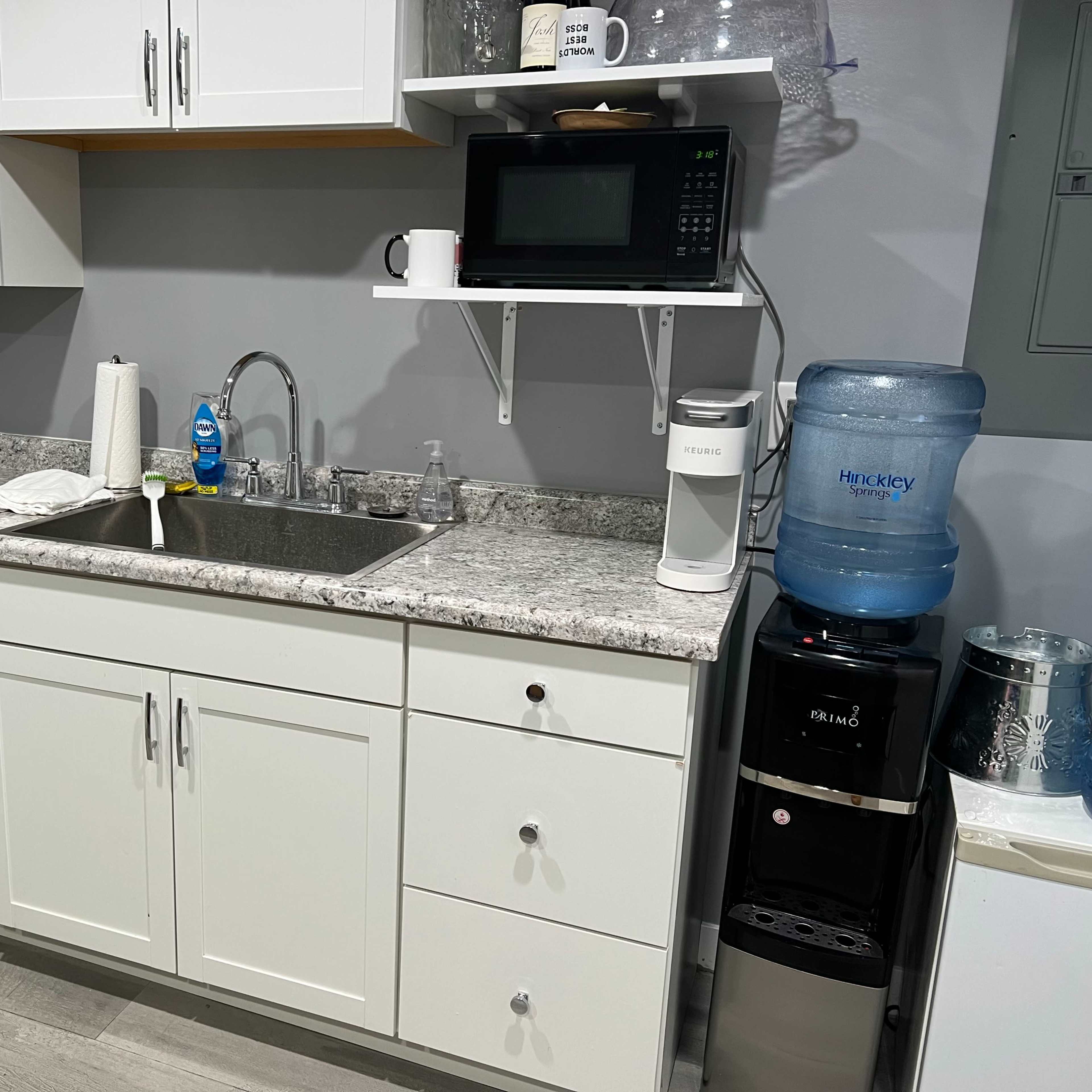 A kitchen sink area with cabinets, a countertop, a microwave, a water dispenser, and a small refrigerator.