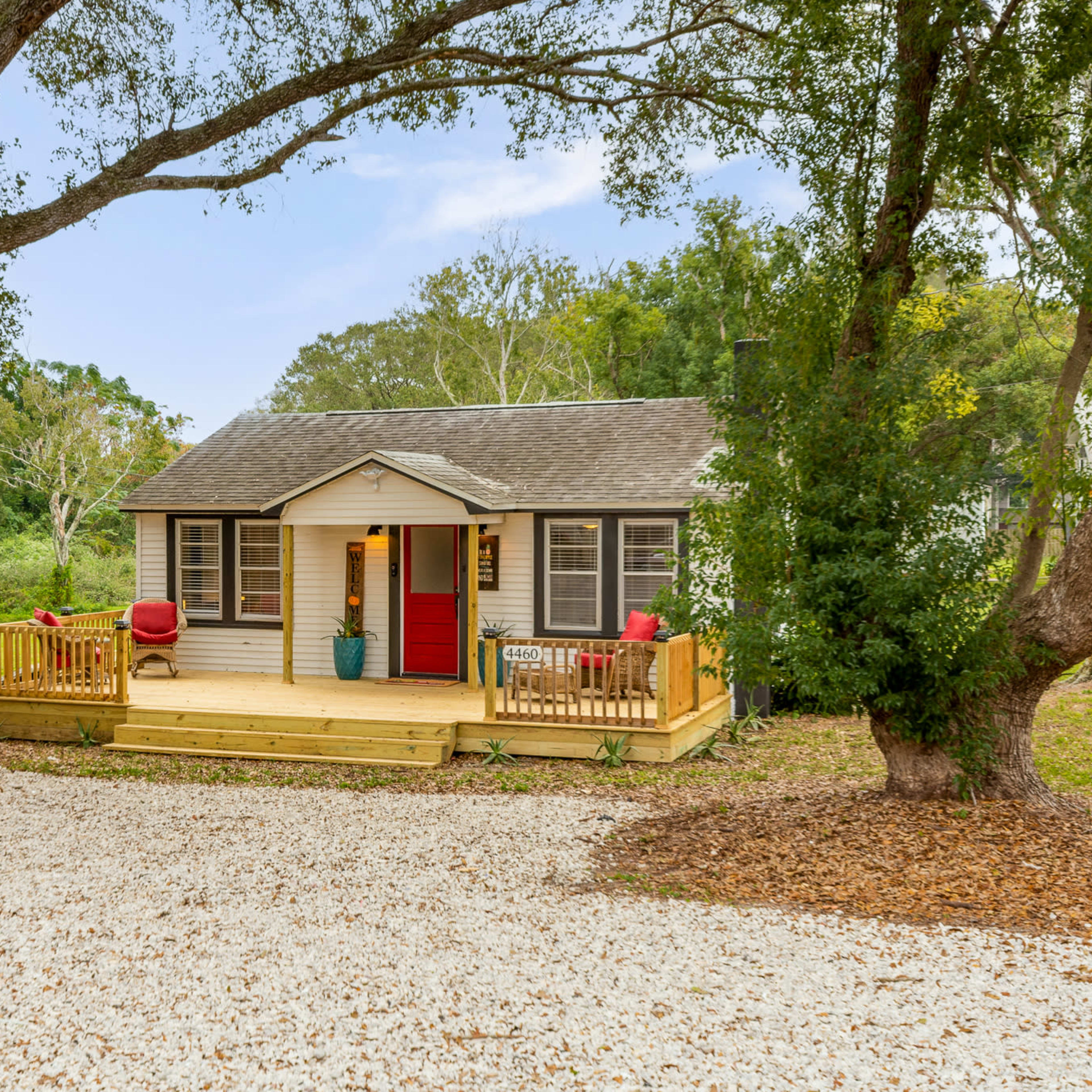 A small, single-story house with a red door and a front porch is situated in a grassy area surrounded by trees.