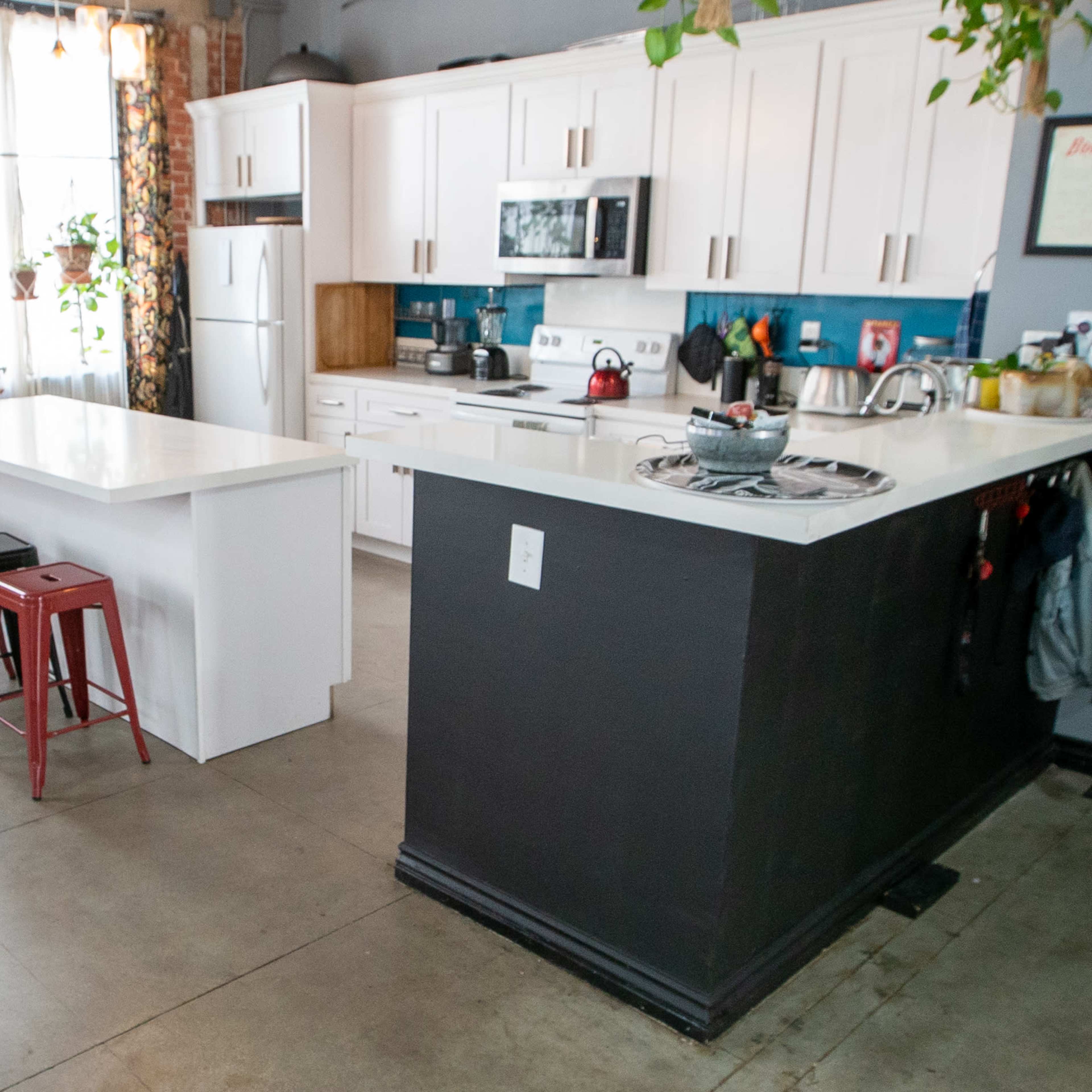 A modern kitchen features a white island with three metal stools, surrounded by brick walls and large windows with plants.