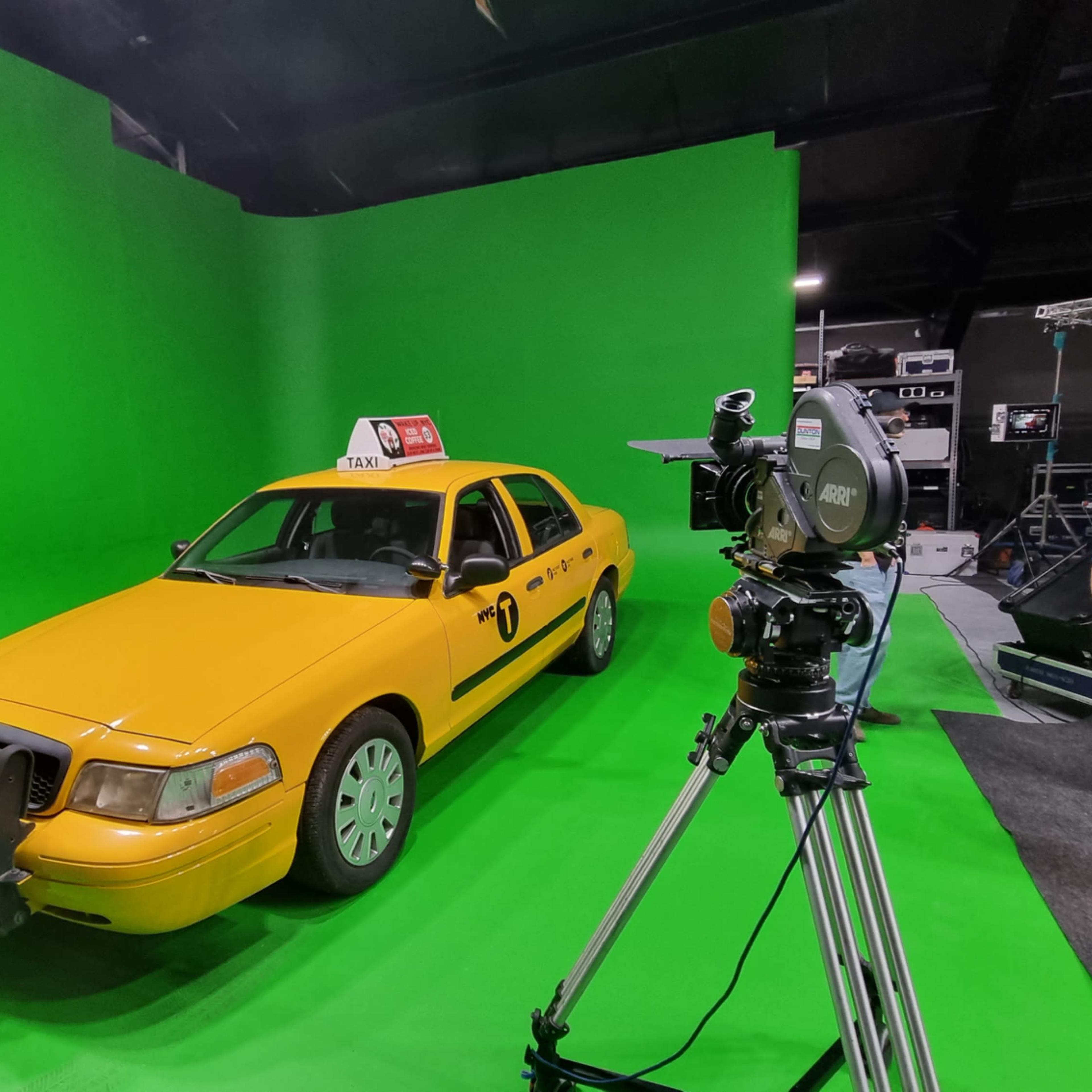 A yellow taxi cab is positioned in front of a green screen in a studio setting, with a camera setup nearby.