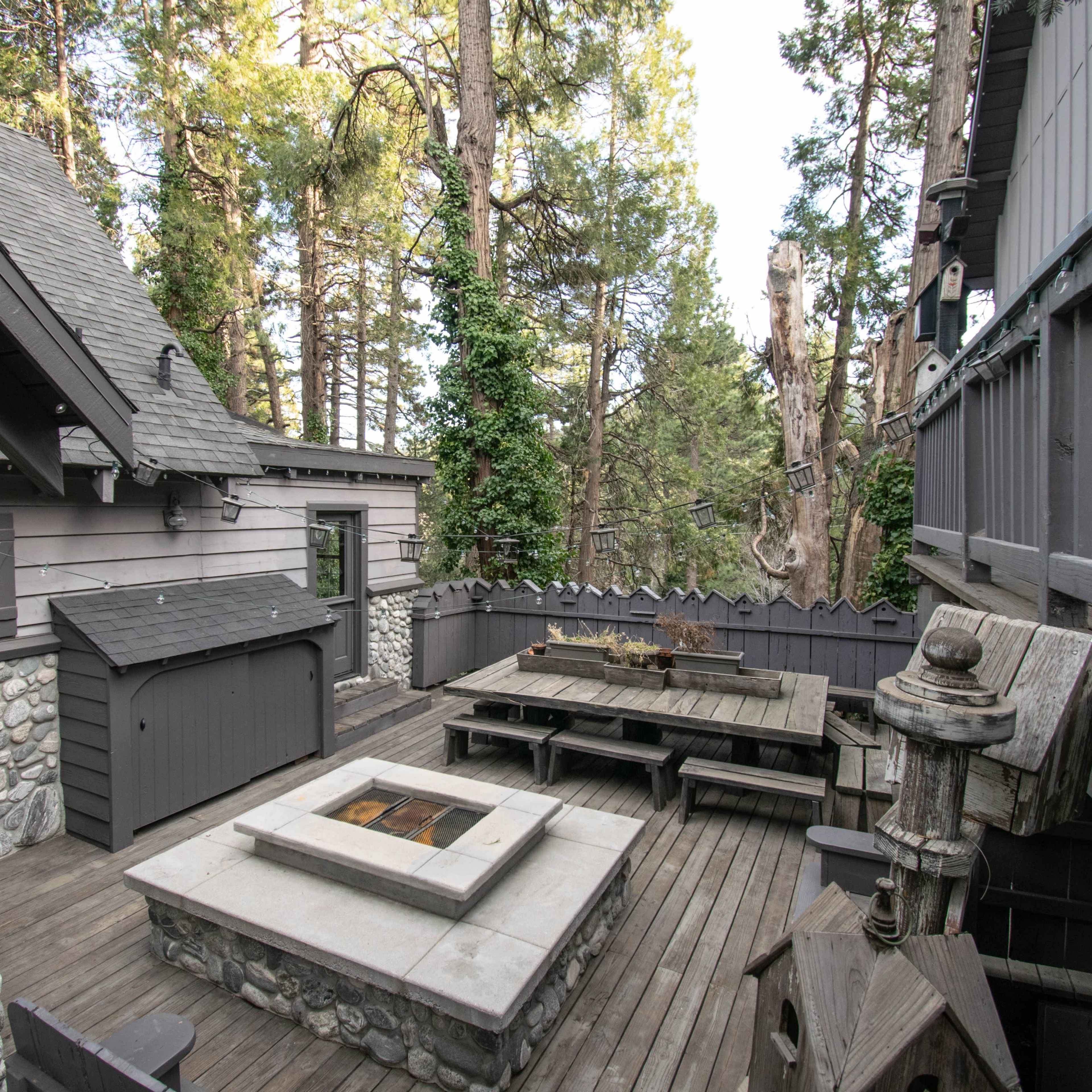 A wooden deck area adjacent to a house, featuring a stone fire pit and a dining table surrounded by tall trees.