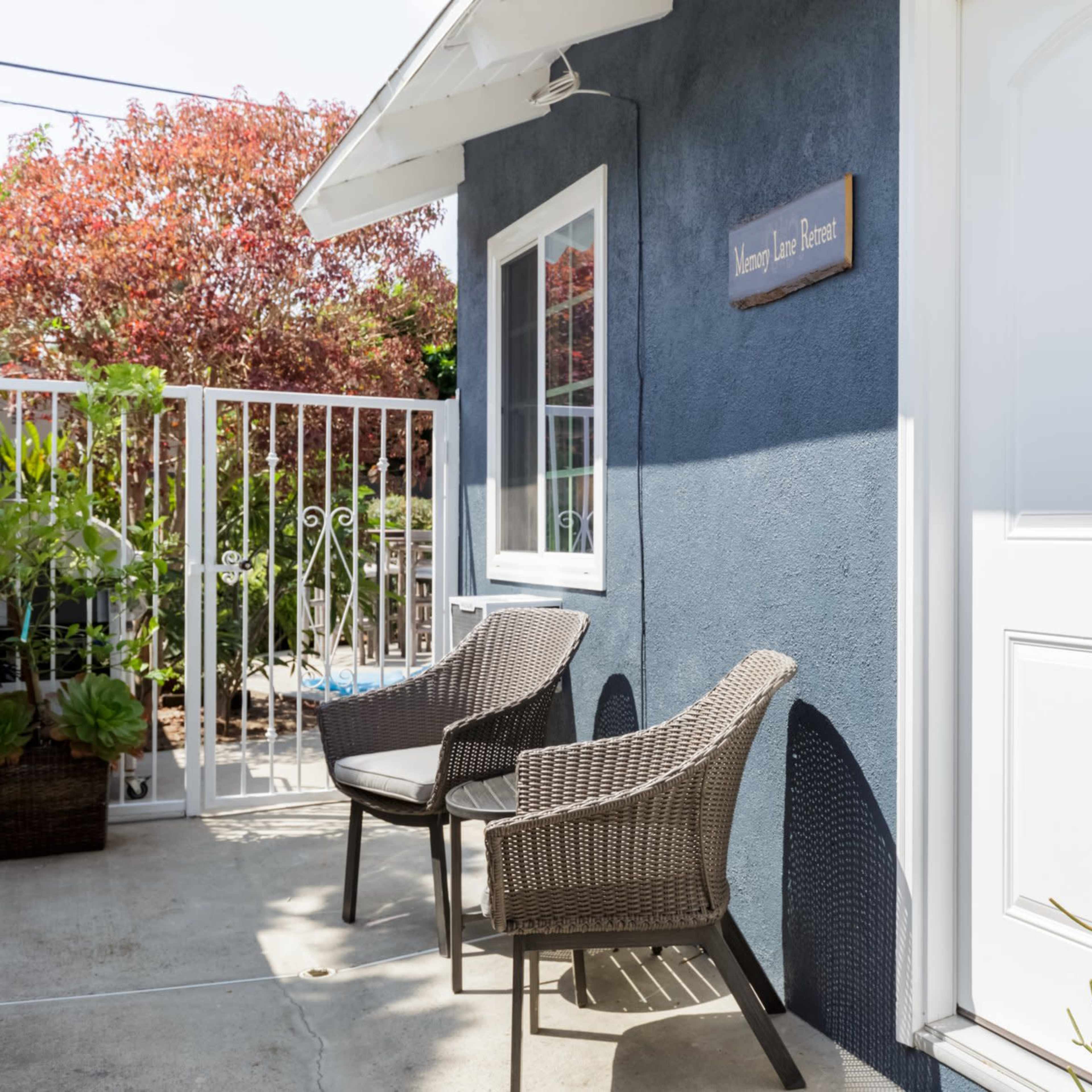 A blue house with a white door features two brown wicker chairs on a concrete patio beside a sign that reads "Journey Lane House."