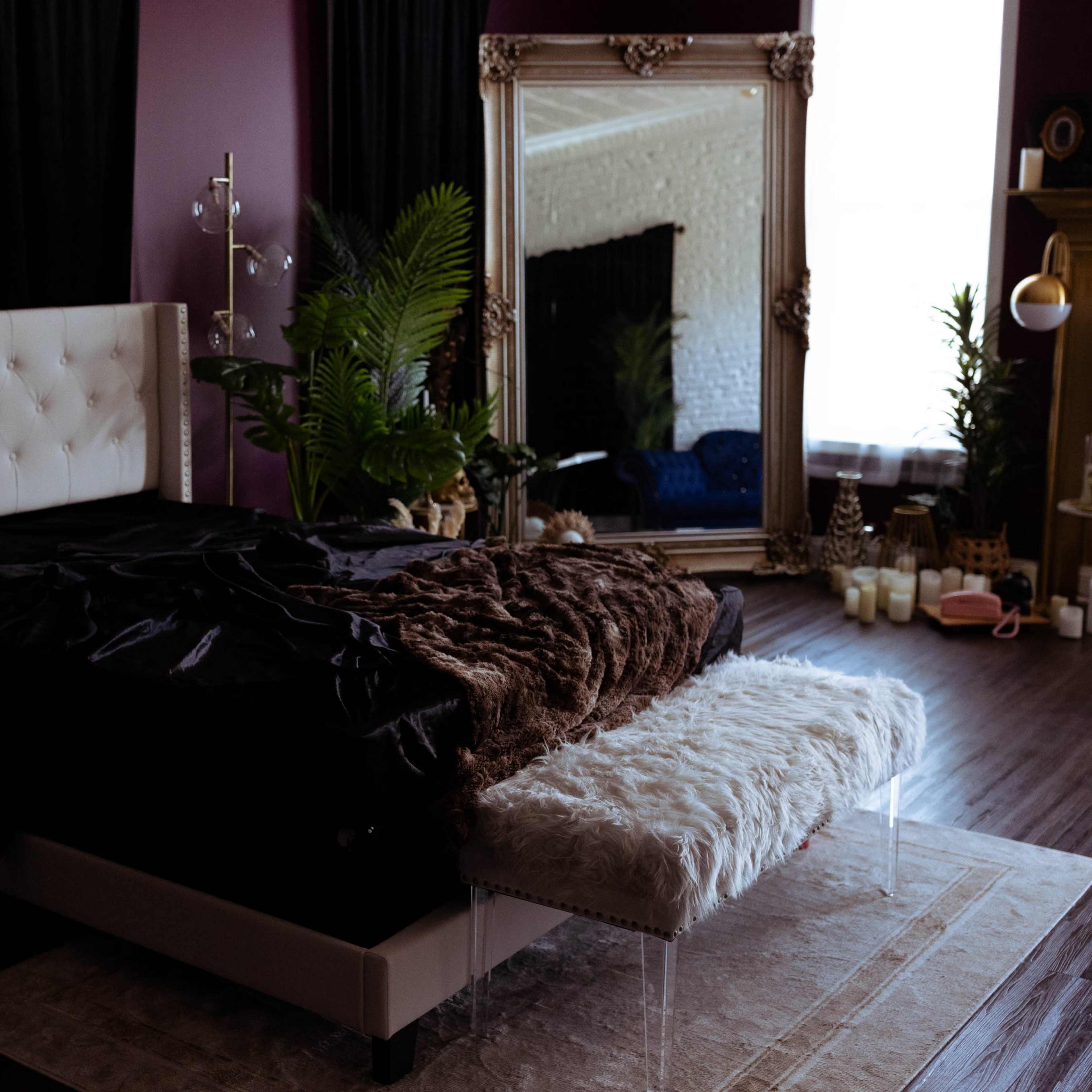 A modern bedroom features a large upholstered bed with a dark and light throw blanket, surrounded by plants and a decorative mirror.