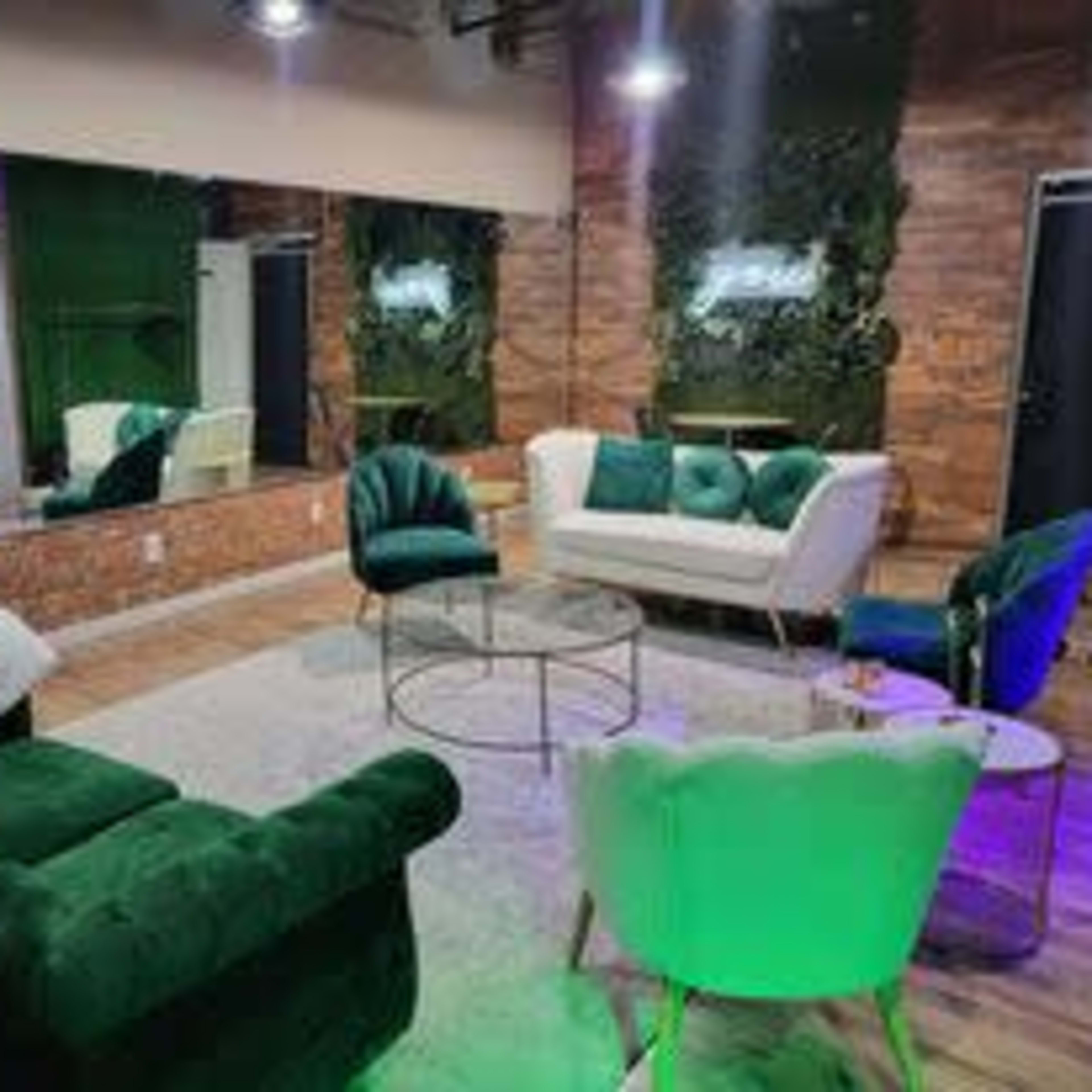 A modern lounge area with green velvet chairs, a white sofa, a circular glass table, and a large mirror in a room with greenery accents.