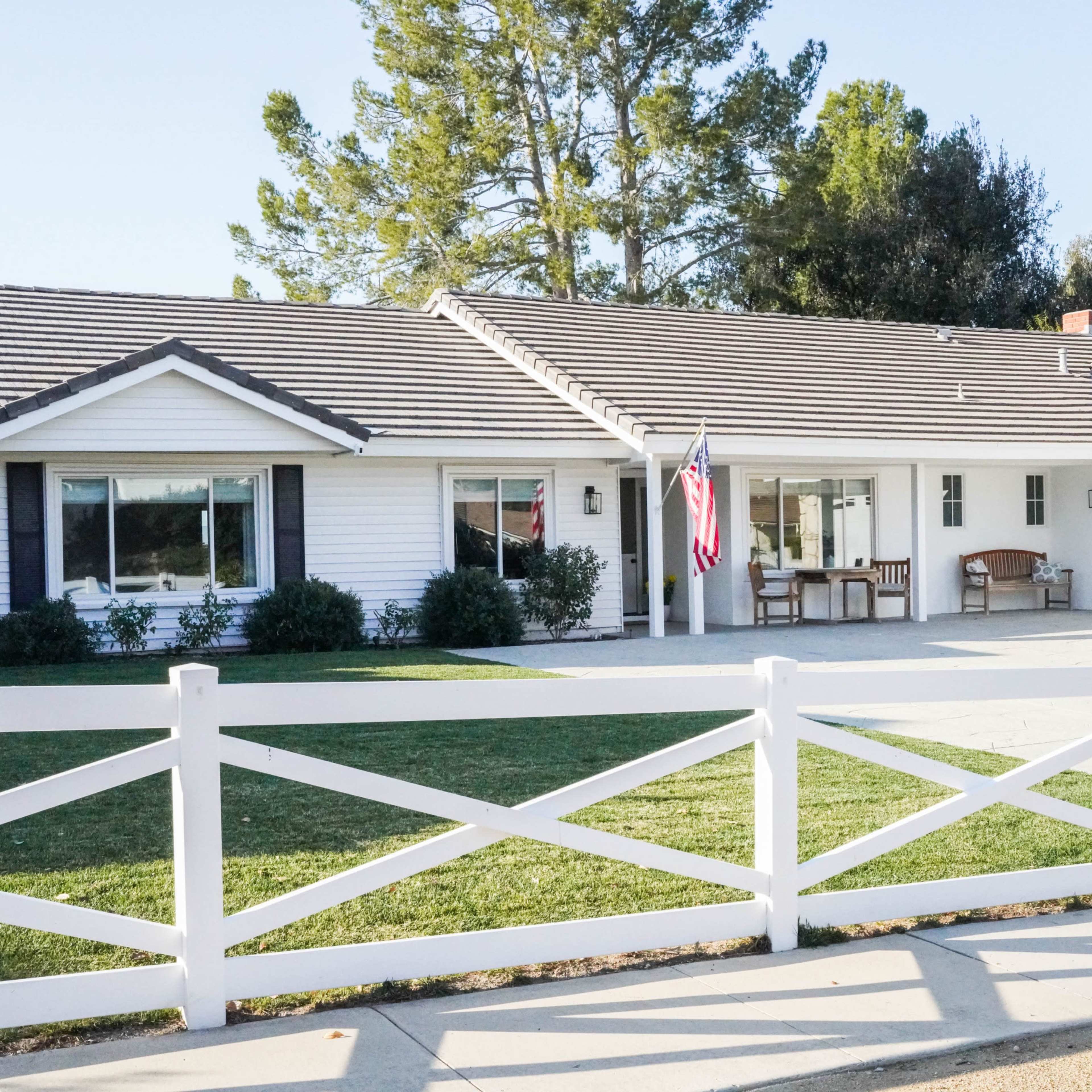 A single-story house with white siding and a grey roof features a flagpole with an American flag in the front yard, surrounded by a white fence.